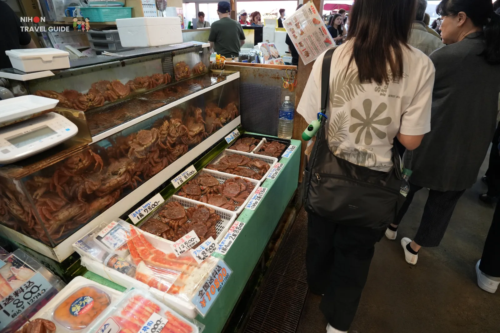 Visitors standing beside stacked tanks of live crabs and trays of packaged seafood inside Hakodate Morning Market.