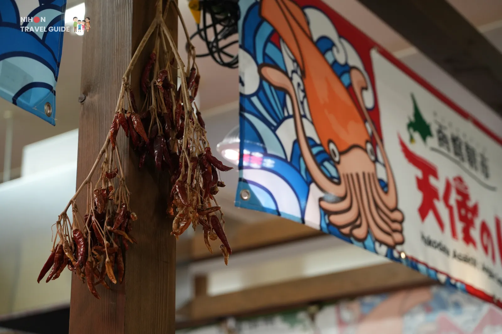 Dried red chili peppers hanging from a wooden post beside a colorful squid banner inside Hakodate Morning Market.