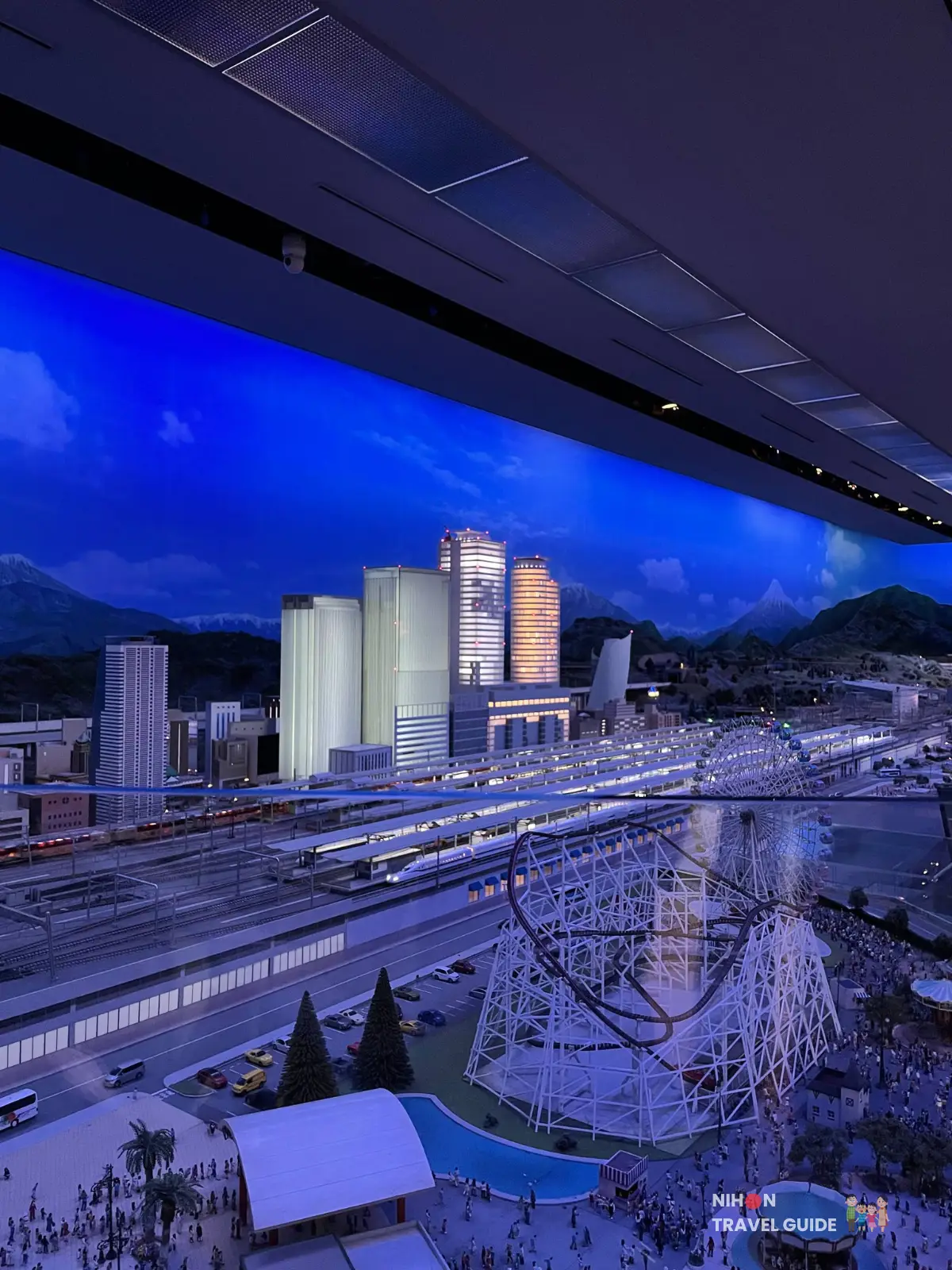 Wide view of the SCMaglev and Railway Park city diorama at night, showing illuminated skyscrapers, multiple train tracks, a roller coaster, and crowds in front of the station.