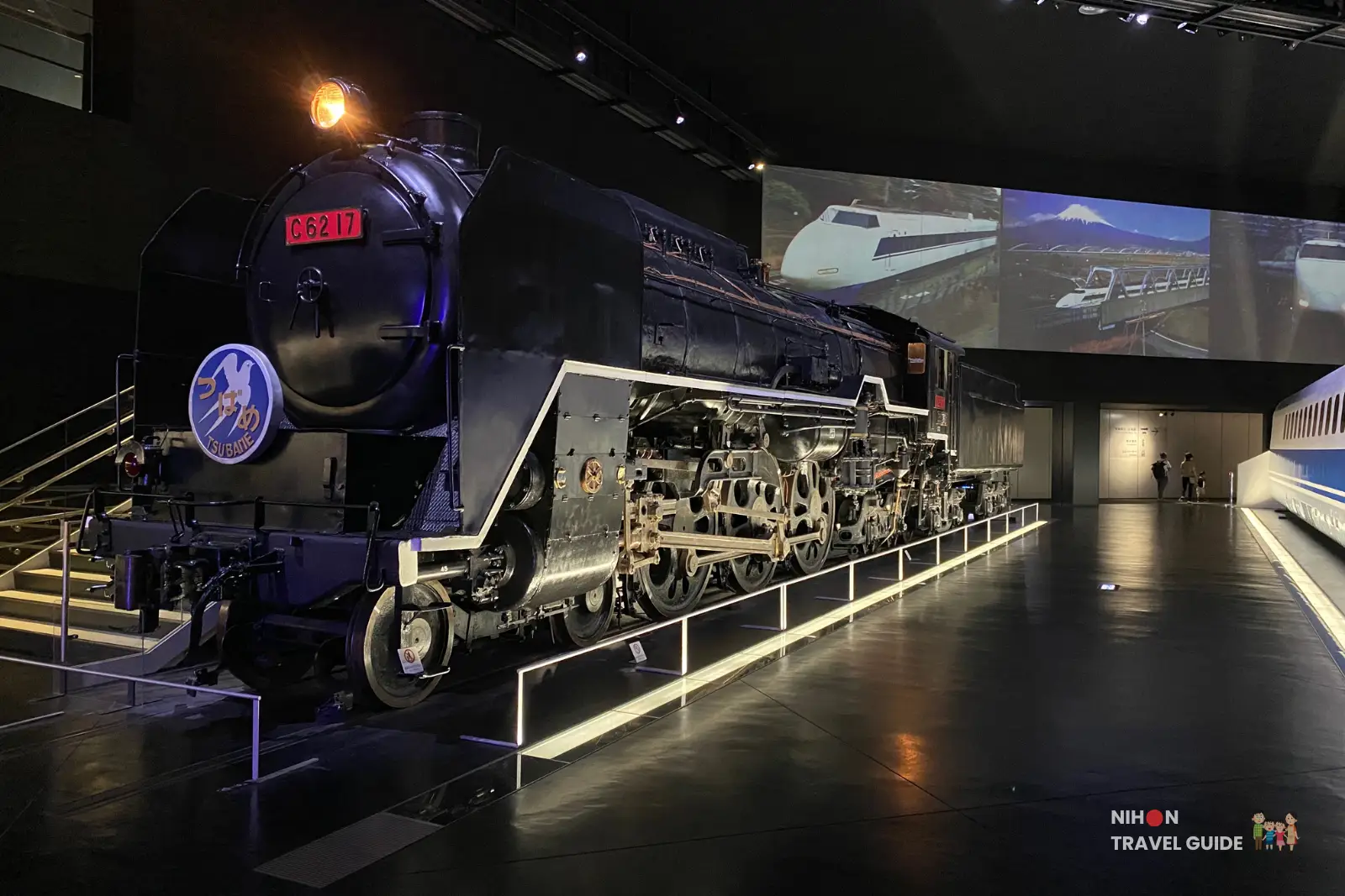 Black C62 steam locomotive on display indoors at SCMaglev and Railway Park, with illuminated wheels and historical train images projected on the wall behind it.