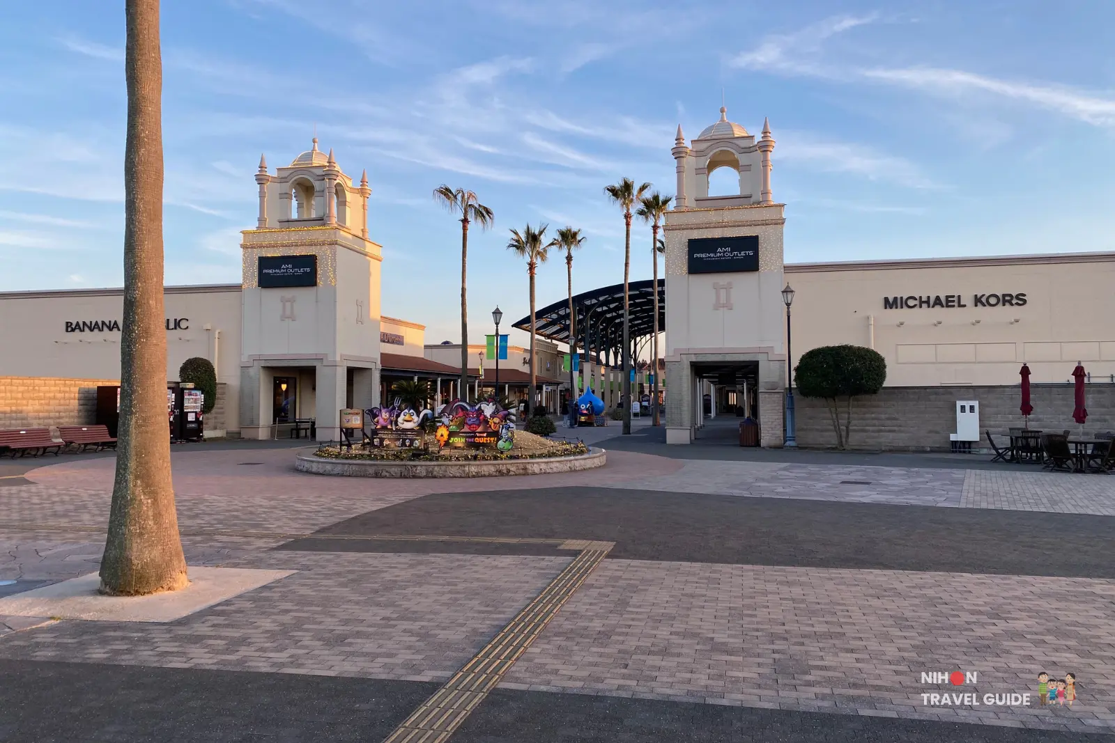 Entrance area of Ami Premium Outlets near Tsukuba with palm trees, brand stores, and open plaza at sunset.