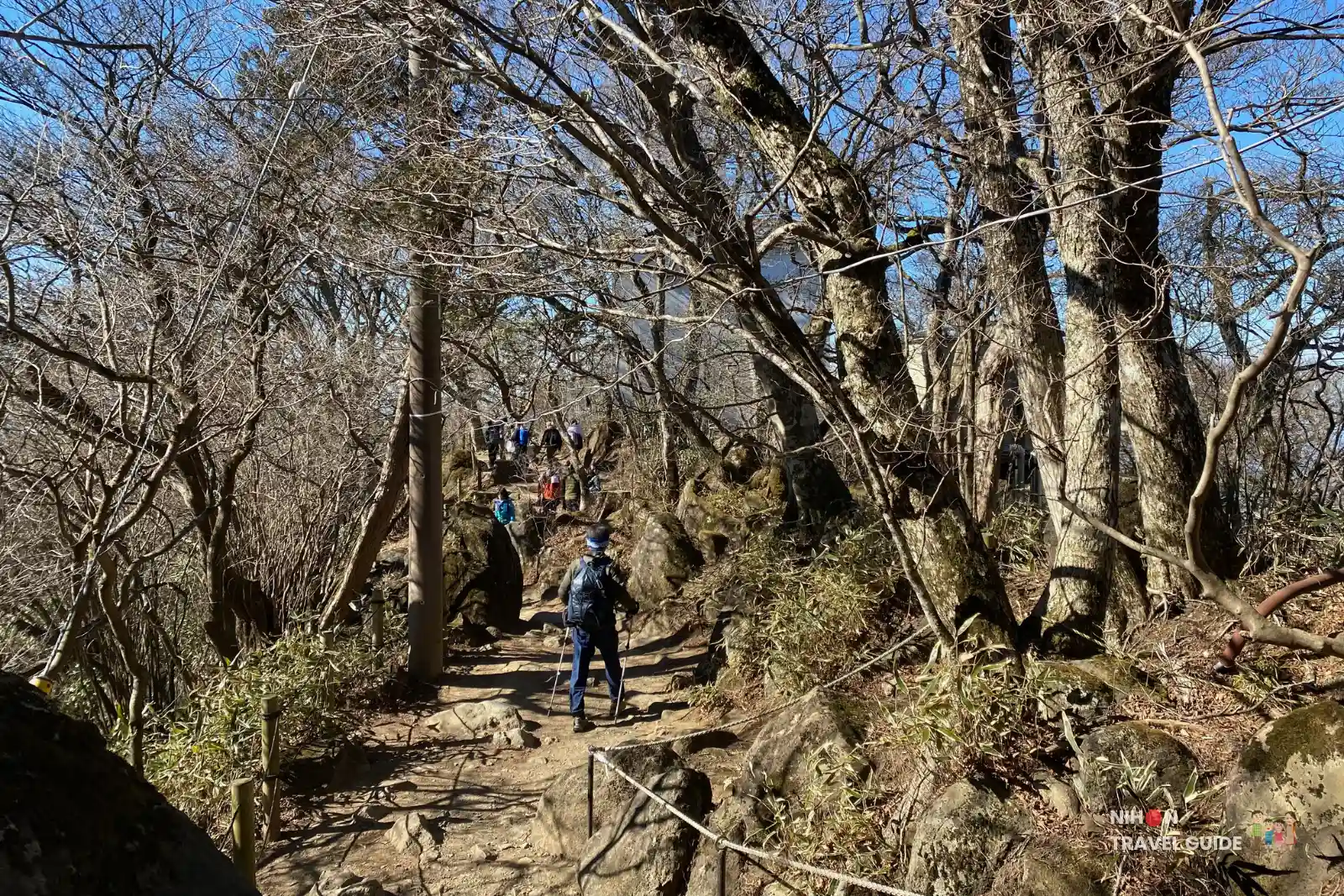 Hikers making their way up the rocky trail between the peaks of Mount Tsukuba, surrounded by bare winter trees.