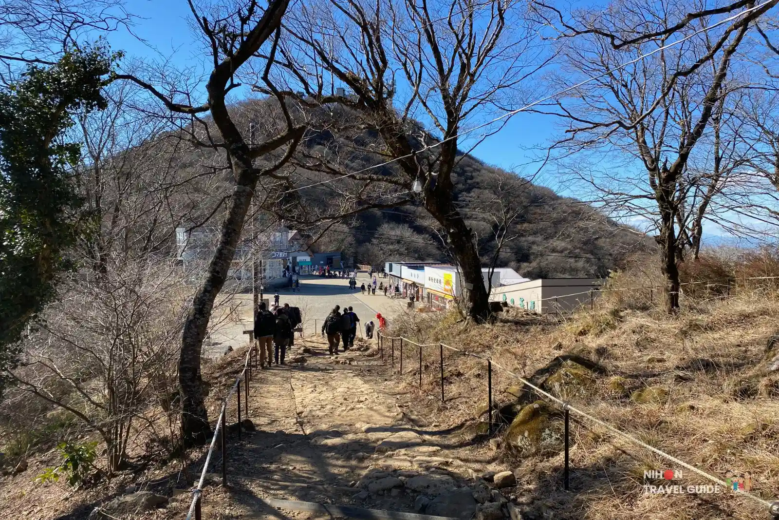 Hikers descending stone steps towards the Miyukigahara Summit Village on Mount Tsukuba, with shops and the Koma Observation Deck visible ahead.