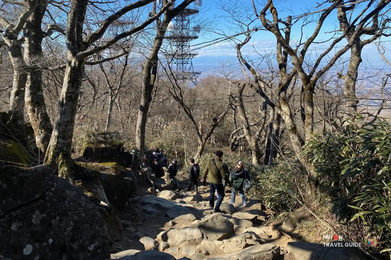 Hikers descending the boulder-strewn path from Nantaisan Peak on Mount Tsukuba with the observation tower and Kanto Plain visible in the background. 