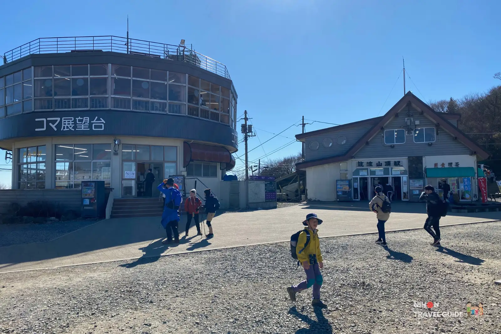 mt-tsukuba-koma-observation-deck Hikers walking near the Koma Observation Deck on Mount Tsukuba, with the round glass-walled observation building on the left and the Tsukuba summit station building on the right under a clear blue sky.
