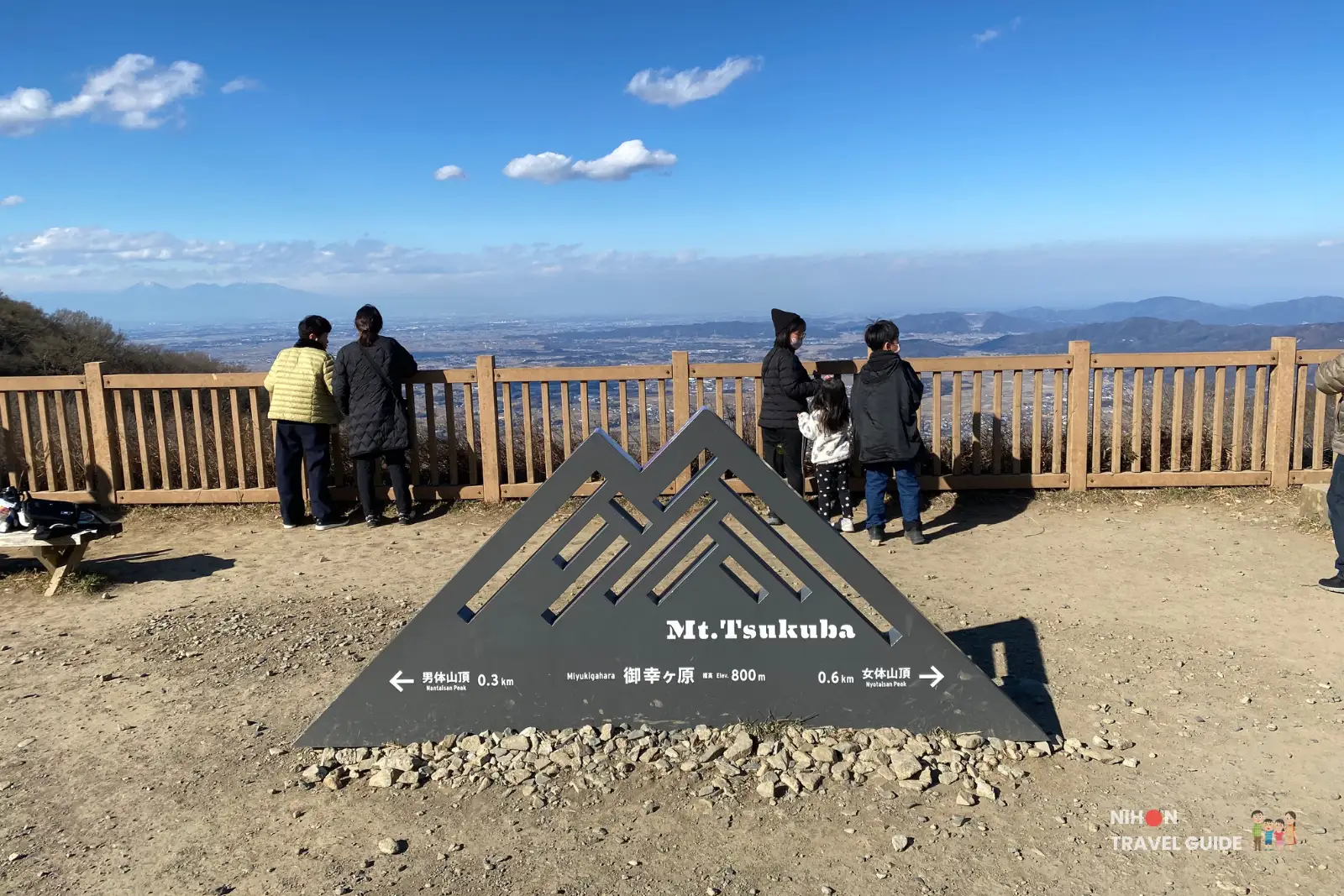mt-tsukuba-miyukigaoka-viewpoint-sign Visitors standing at a wooden fenced viewpoint on Mount Tsukuba, with a triangular “Mt. Tsukuba” direction sign in the foreground and a wide city and mountain panorama under a blue sky.