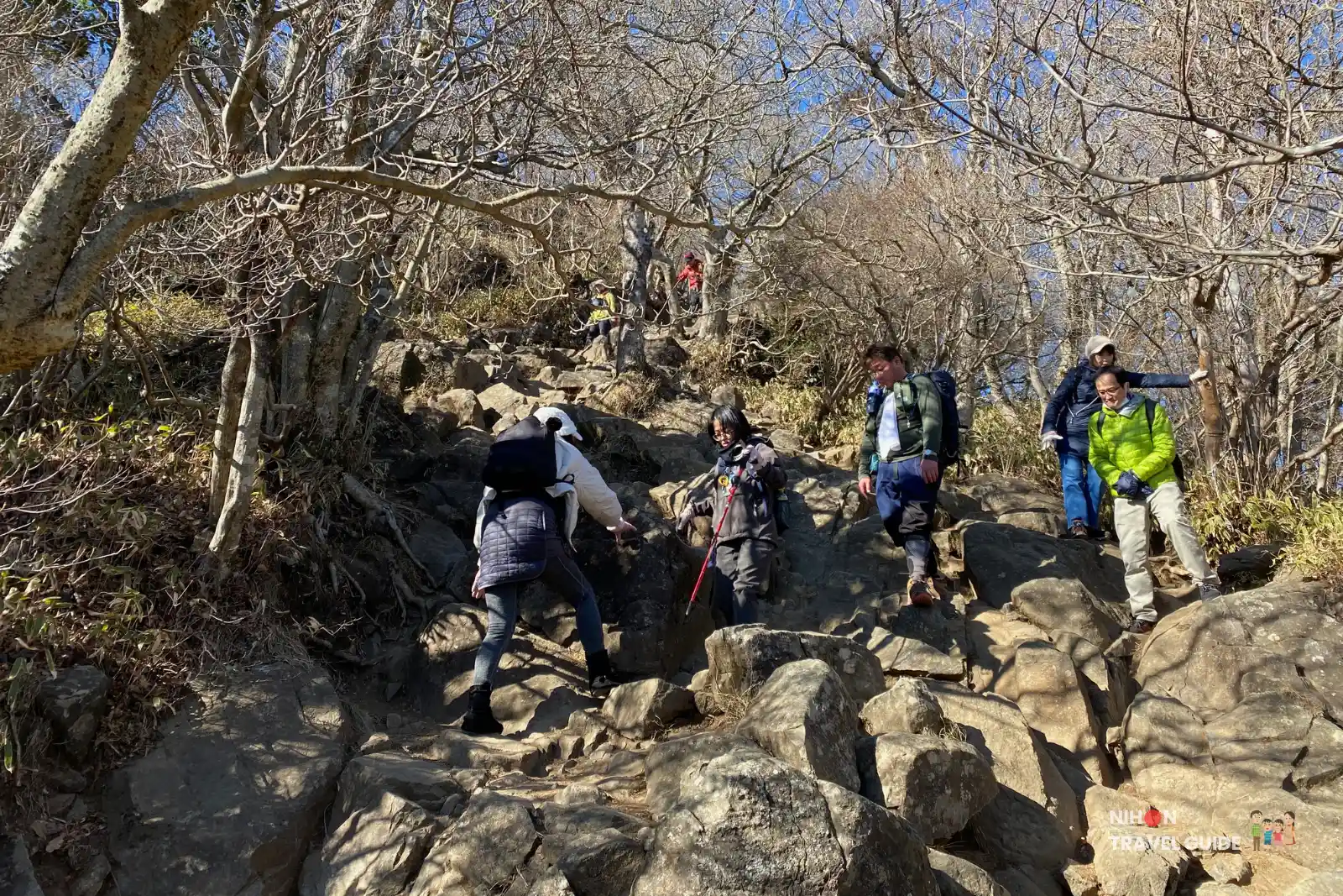 Hikers scrambling up the steep final rocky slope to Nantaisan Peak on Mount Tsukuba, surrounded by leafless trees under a clear blue sky.