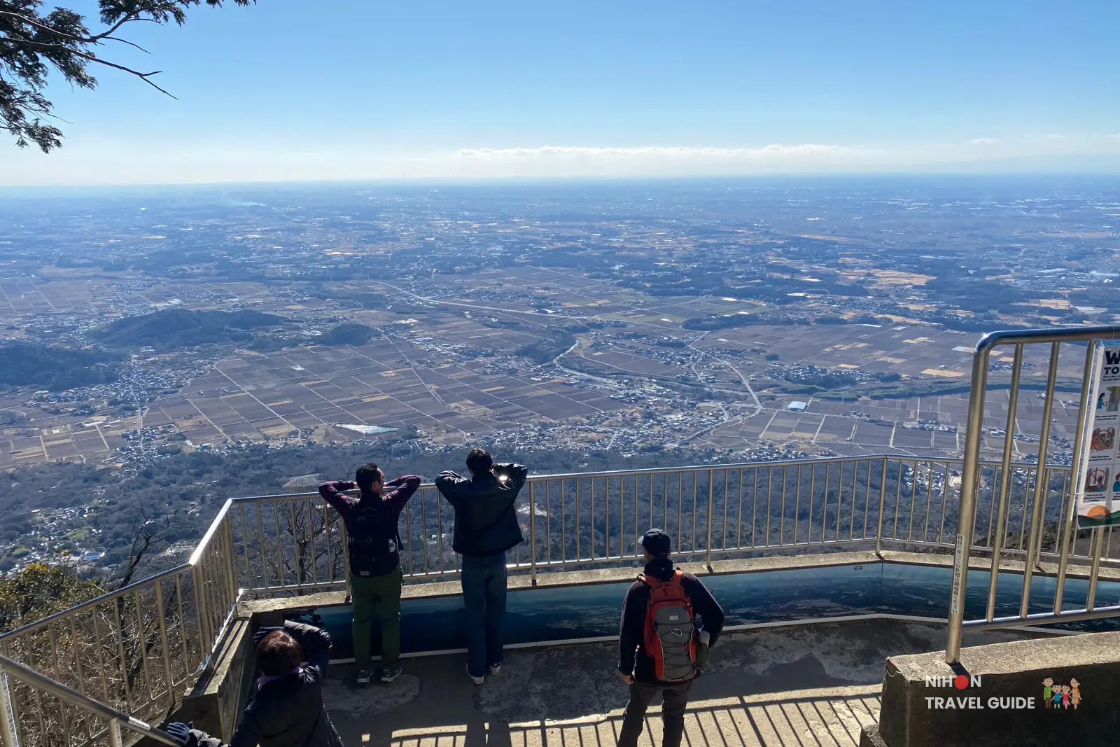 Wide view from Nantaisan Peak on Mount Tsukuba, looking over the Kanto Plain with visitors standing at a fenced observation deck in the lower foreground.