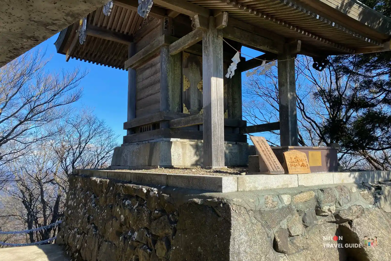 mt-tsukuba-nantaisan-summit-shrine Small wooden summit shrine on Nantaisan Peak at Mount Tsukuba, raised on a stone platform with decorative paper streamers, surrounded by bare trees under a clear blue sky.