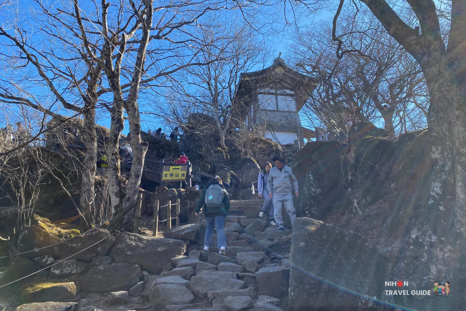 mt-tsukuba-nyotaisan-peak-approach-trail Hikers climbing a rocky stone stairway through leafless trees toward the shrine building near the summit of Nyotaisan Peak on Mount Tsukuba under a clear blue sky.