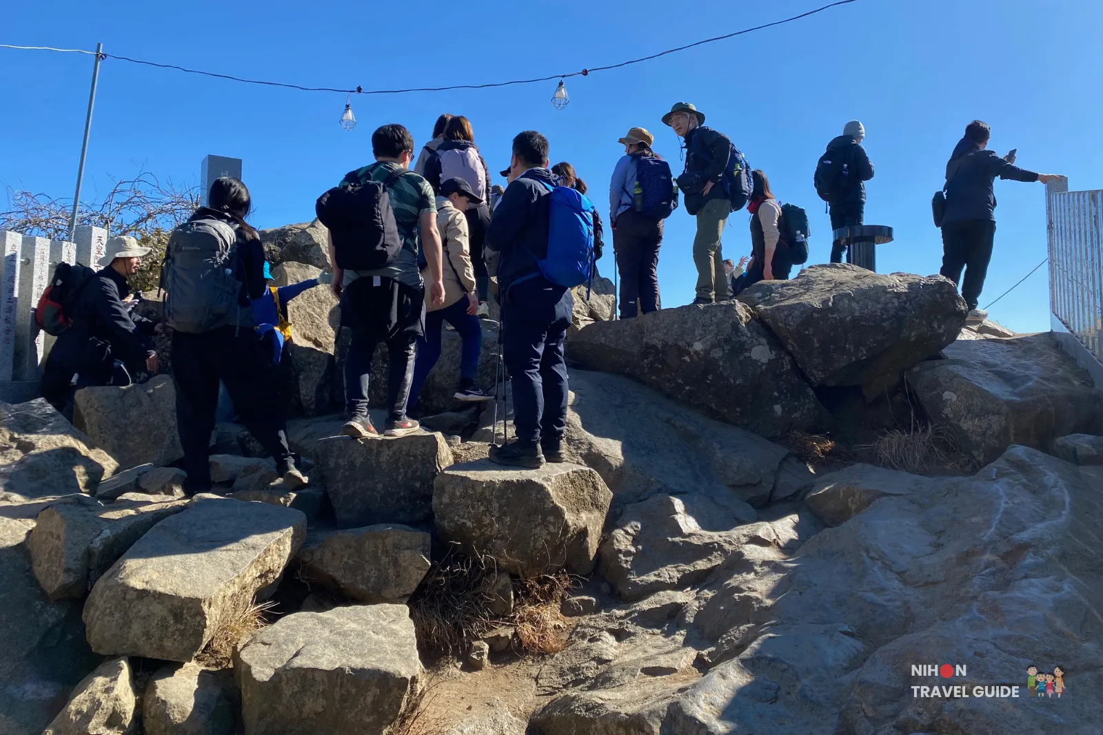 mt-tsukuba-nyotaisan-peak-queue Hikers queuing on rocky steps near the summit of Nyotaisan Peak on Mount Tsukuba, waiting their turn to stand at the top and enjoy the panoramic view under a clear blue sky.