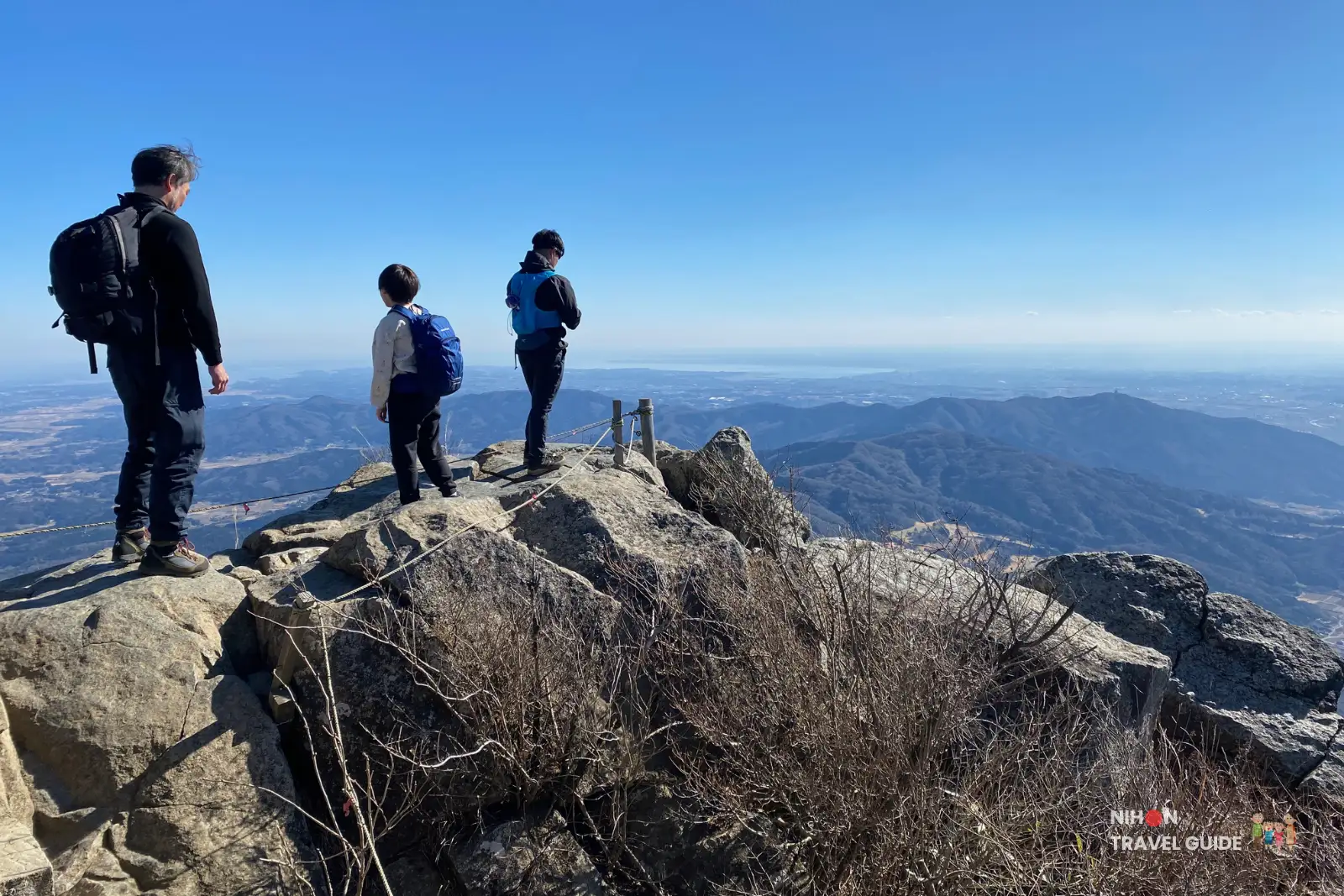 Hikers standing on the rocky summit of Nyotaisan Peak on Mount Tsukuba, looking out over ranges of blue mountains and the distant plains under a clear sky.