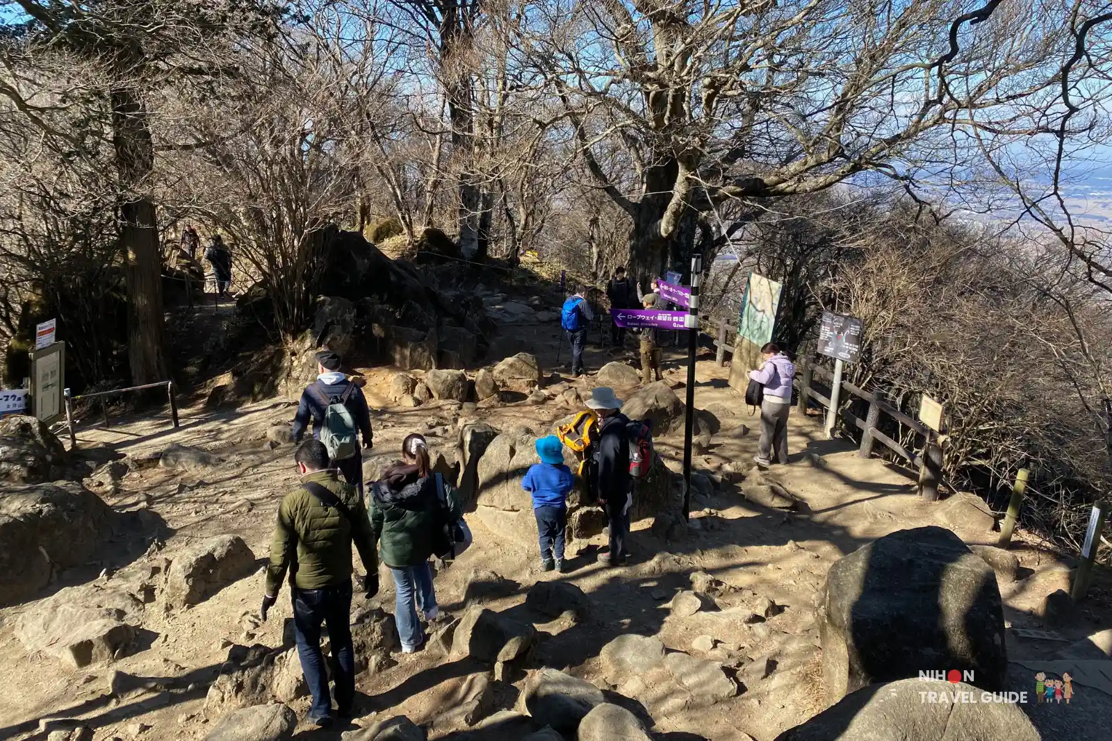 View over the Nyotaisan rest area on Mount Tsukuba, with hikers walking among rocks and trees, purple trail signs, and distant valley scenery visible through the bare branches.