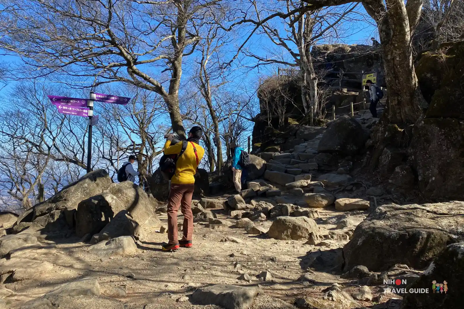 Hikers standing in a rocky clearing at the Nyotaisan rest area on Mount Tsukuba, with purple trail signs and stone steps leading up toward the summit through leafless trees.
