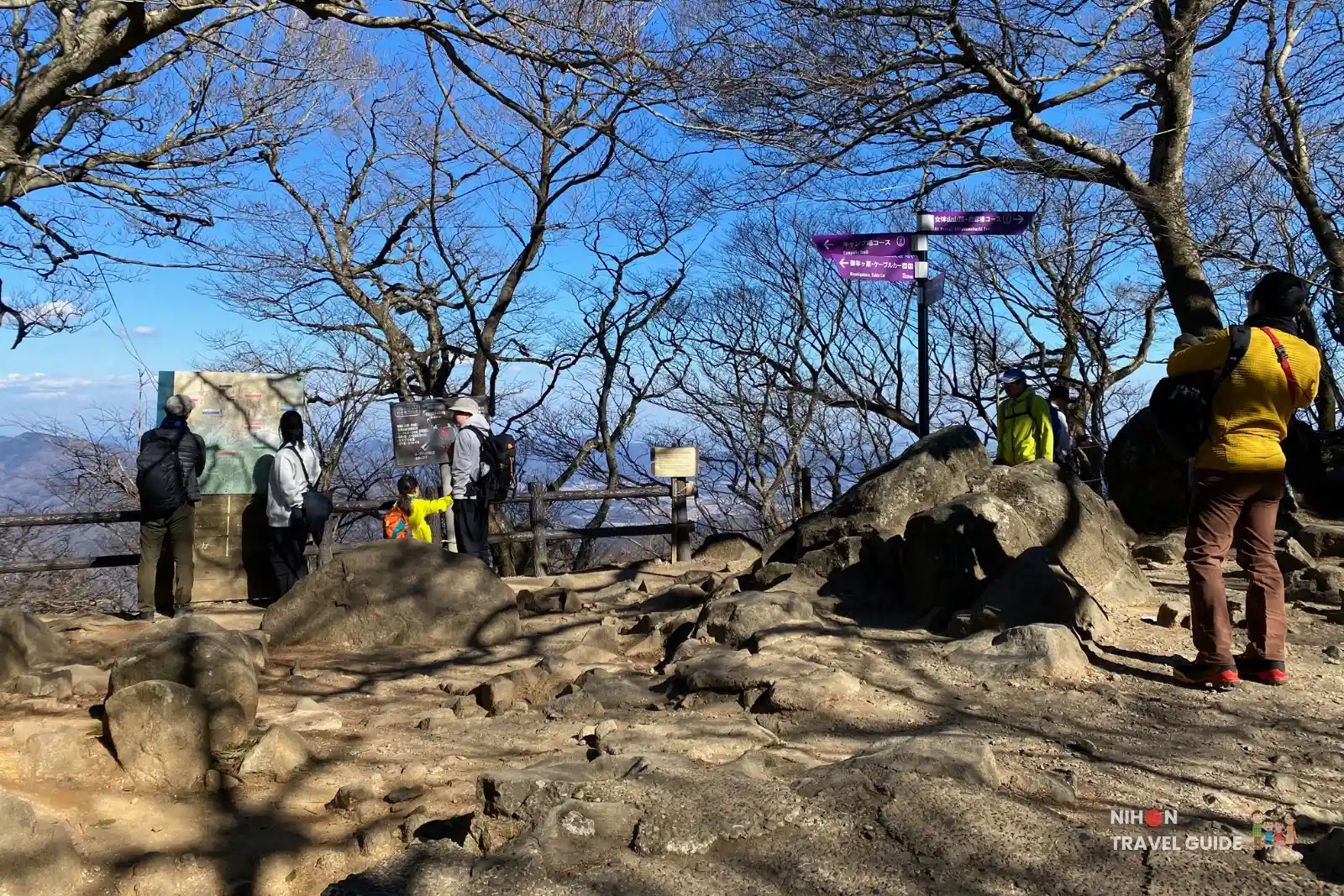Hikers standing at a rocky clearing and trail junction just before the climb to Nyotaisan Peak on Mount Tsukuba, with bare trees, map boards, and purple direction signs under a blue sky.