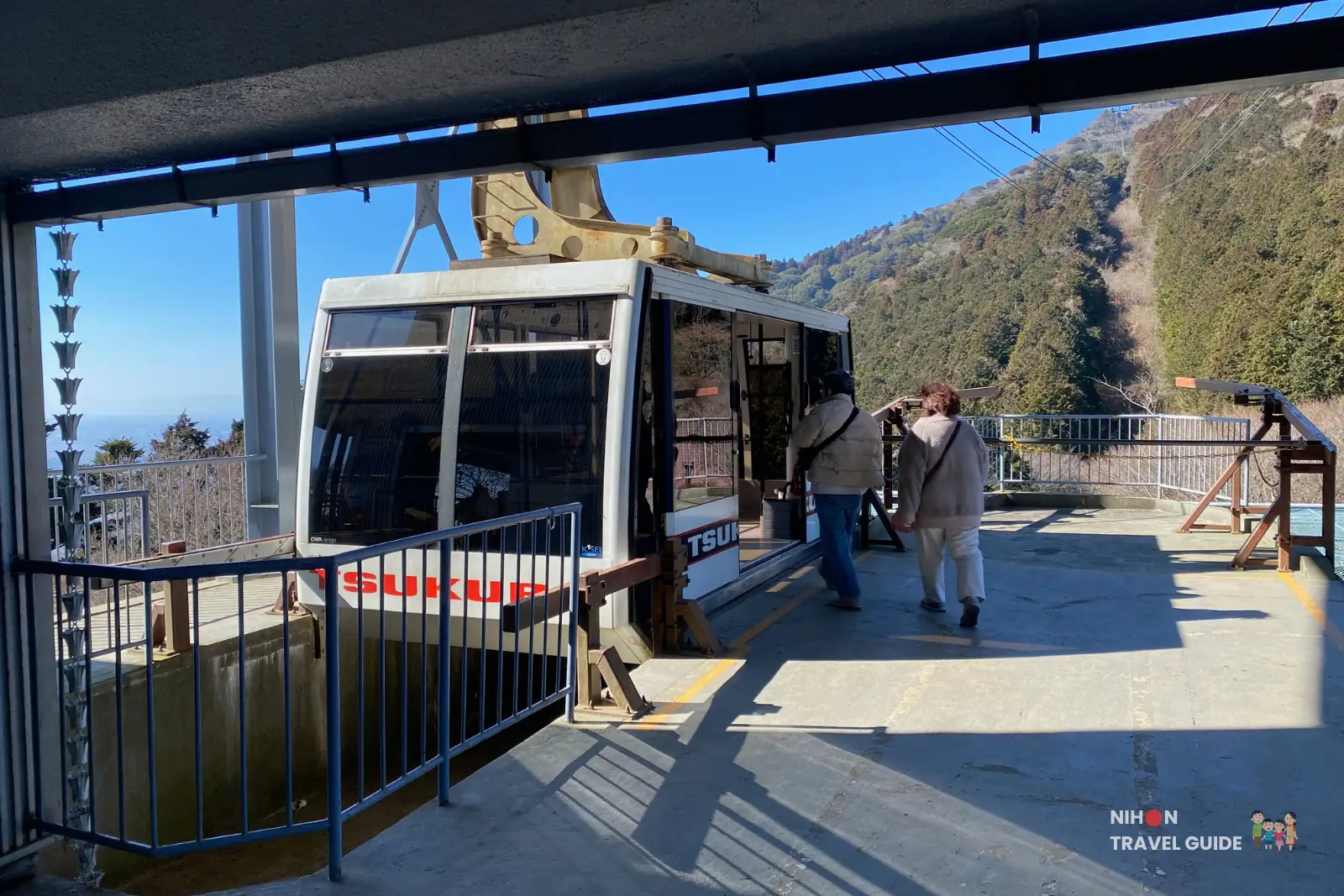 mt-tsukuba-ropeway-cable-car-platform Passengers boarding a Mt. Tsukuba ropeway cable car at the mountain station, with the gondola stopped at the platform and forested slopes visible in the background.