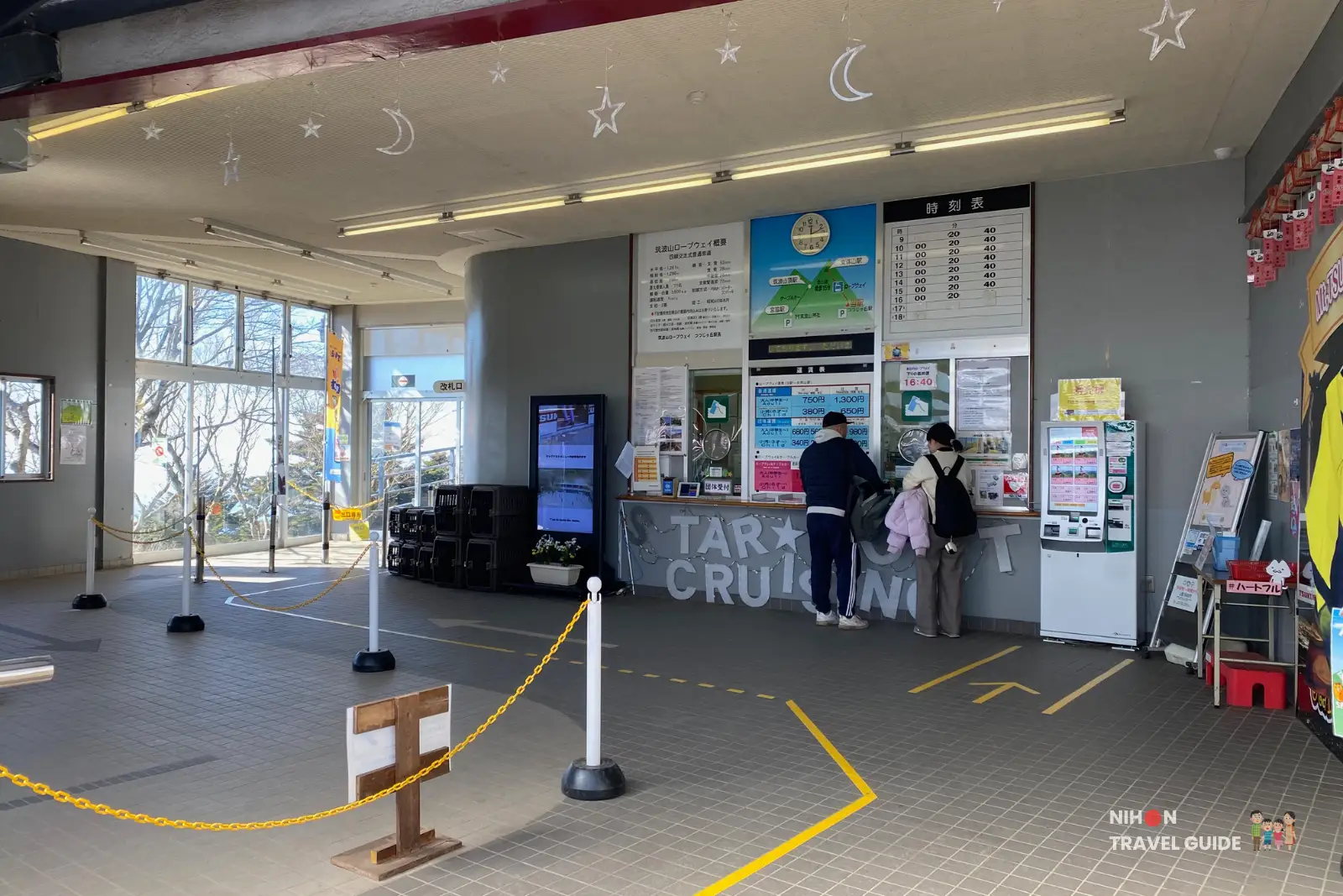 mt-tsukuba-ropeway-ticketing-station Interior of the Mt. Tsukuba ropeway station ticketing hall, with two visitors at the ticket counter, pet cages stacked near a digital information screen, and queuing lines marked on the tiled floor.