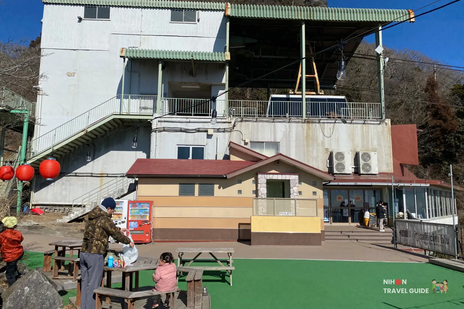 mt-tsukuba-summit-ropeway-station Summit ropeway station on Mount Tsukuba, with the multi-level station building behind a small toilet block, picnic tables, red lanterns, and families relaxing in the sunshine.