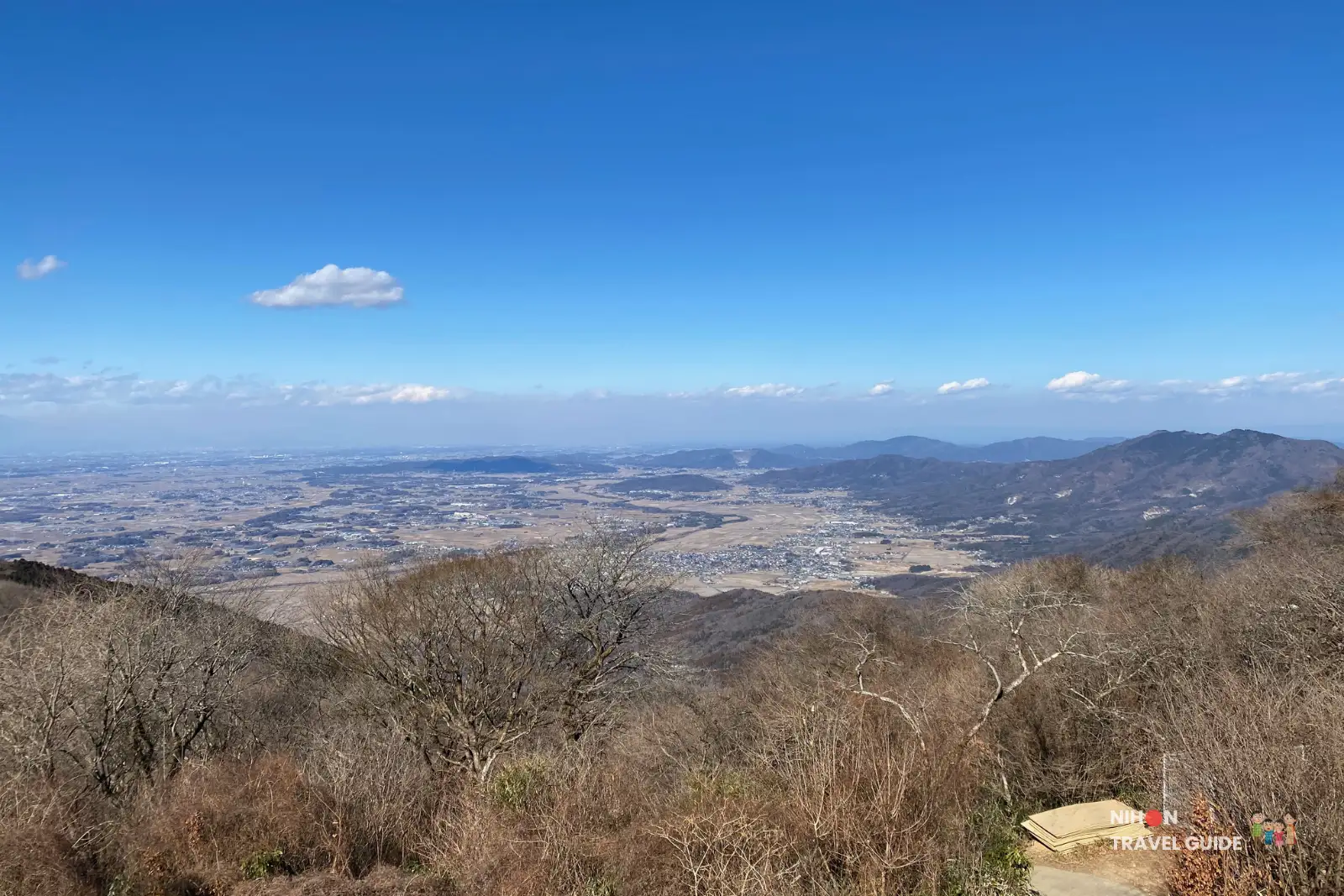 mt-tsukuba-summit-village-view-over-kanto-plain Wide panorama from Mount Tsukuba’s summit village, overlooking the Kanto Plain and surrounding hills under a clear blue sky with a few scattered clouds and leafless trees in the foreground.