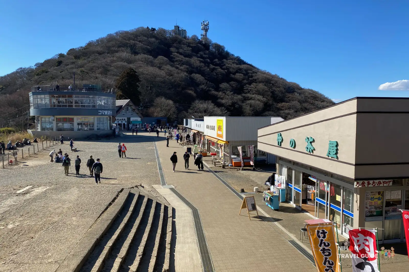 mt-tsukuba-summit-village View over Summit Village on Mount Tsukuba, showing the Koma Observation Deck, a row of small restaurants and souvenir shops, and hikers walking across the open plaza below the wooded peak.