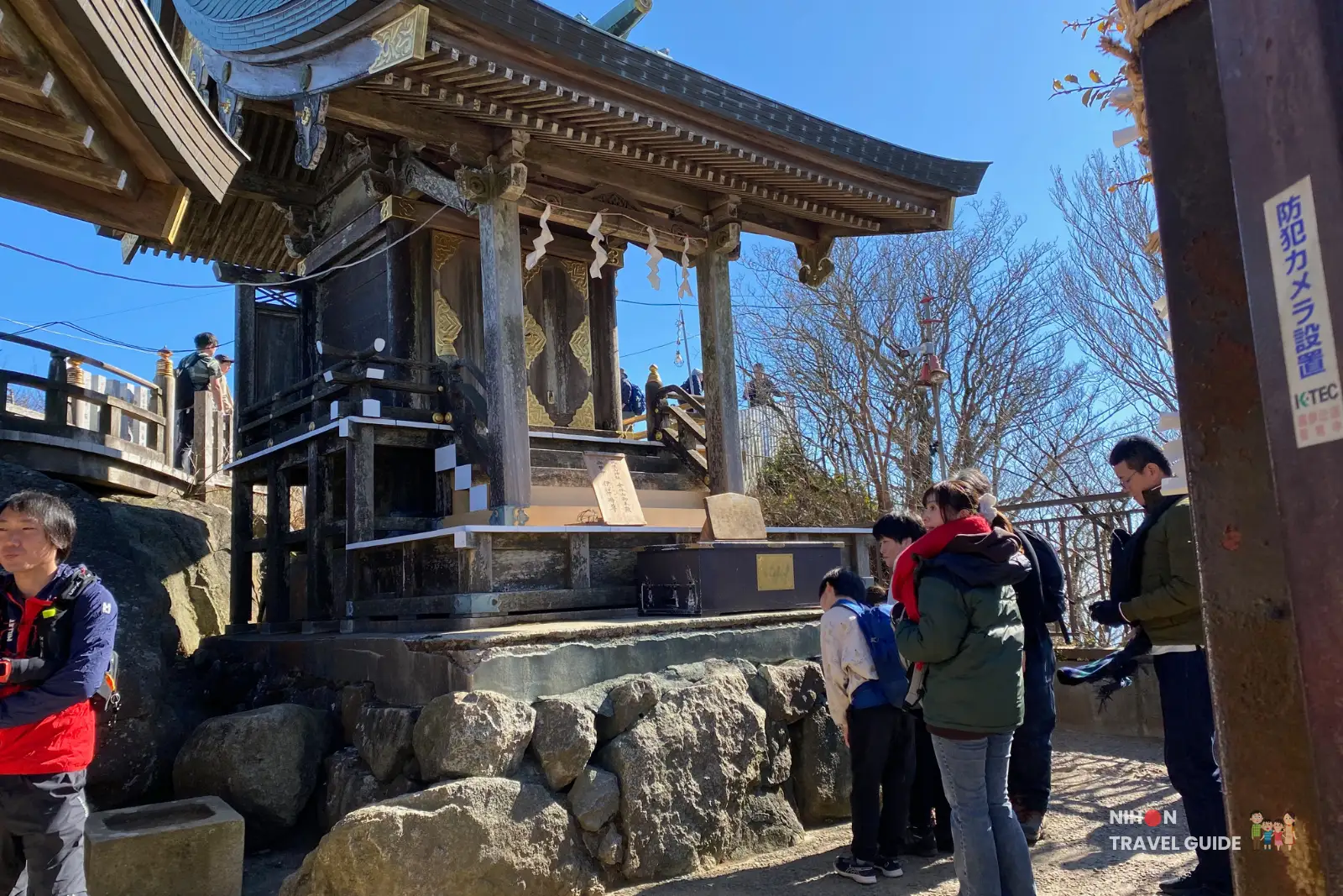 mt-tsukuba-tsukuba-shrine-nyotaisan-peak Visitors praying at the Tsukuba Shrine building on Nyotaisan Peak, a small wooden shrine raised on a stone base with decorative gold details and paper streamers under a clear blue sky.