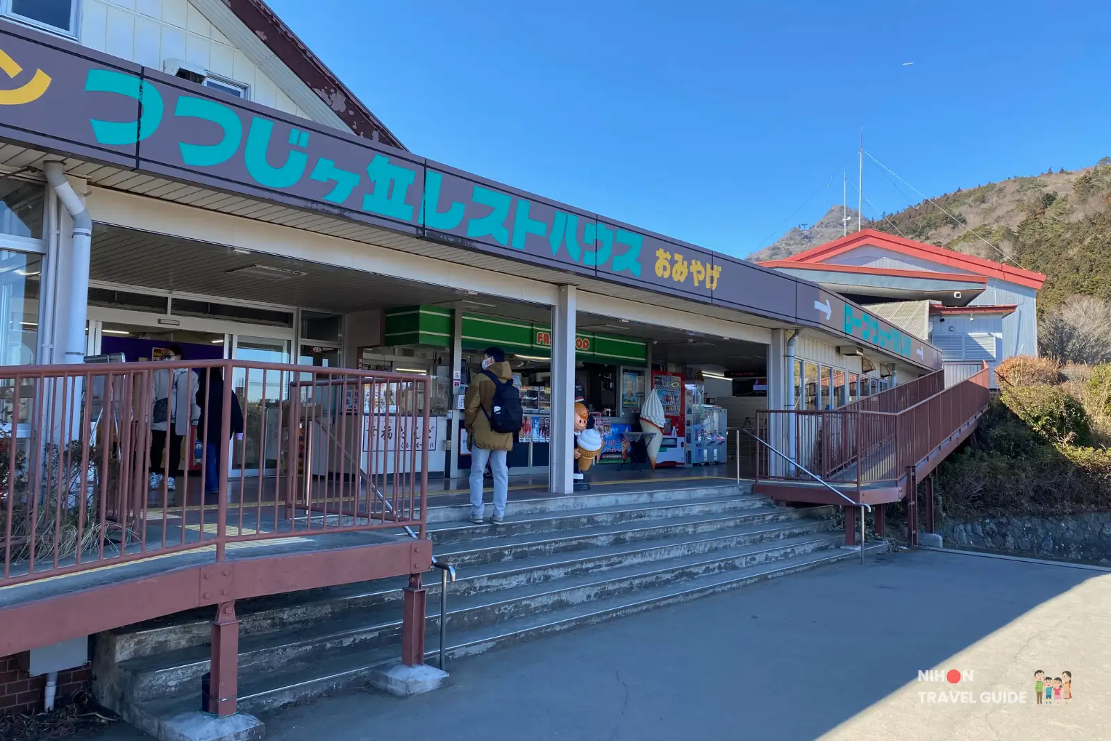 mt-tsukuba-tsutsujigaoka-rest-house Entrance of Tsutsujigaoka Rest House at the base of Mount Tsukuba, with wide steps leading up to the souvenir shop and restaurant under a long signboard and clear blue sky.