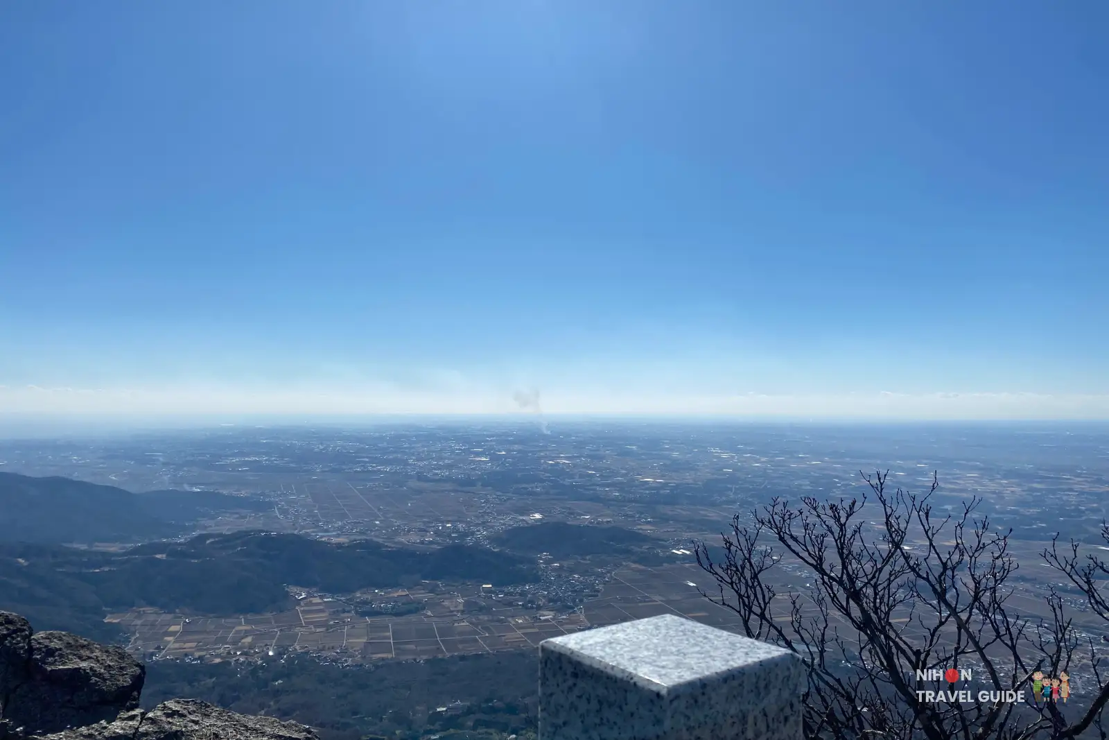 Sweeping view from Nyotaisan Peak on Mount Tsukuba, looking over distant towns and patchwork fields under a clear blue sky, with a granite summit marker and bare branches in the foreground.