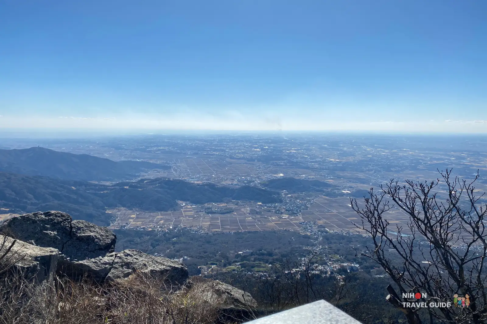 Elevated panorama from Nyotaisan Peak on Mount Tsukuba, showing rocky foreground slopes and the wide Kanto Plain stretching to the hazy horizon beneath a bright blue sky.