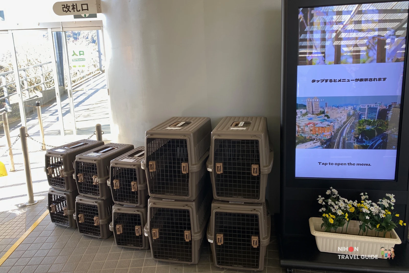 pet-cages-mt-tsukuba-ropeway-base-station Stacked plastic pet cages lined up inside the Mount Tsukuba ropeway base station, next to a large digital information screen and a planter of flowers near the glass entrance doors.
