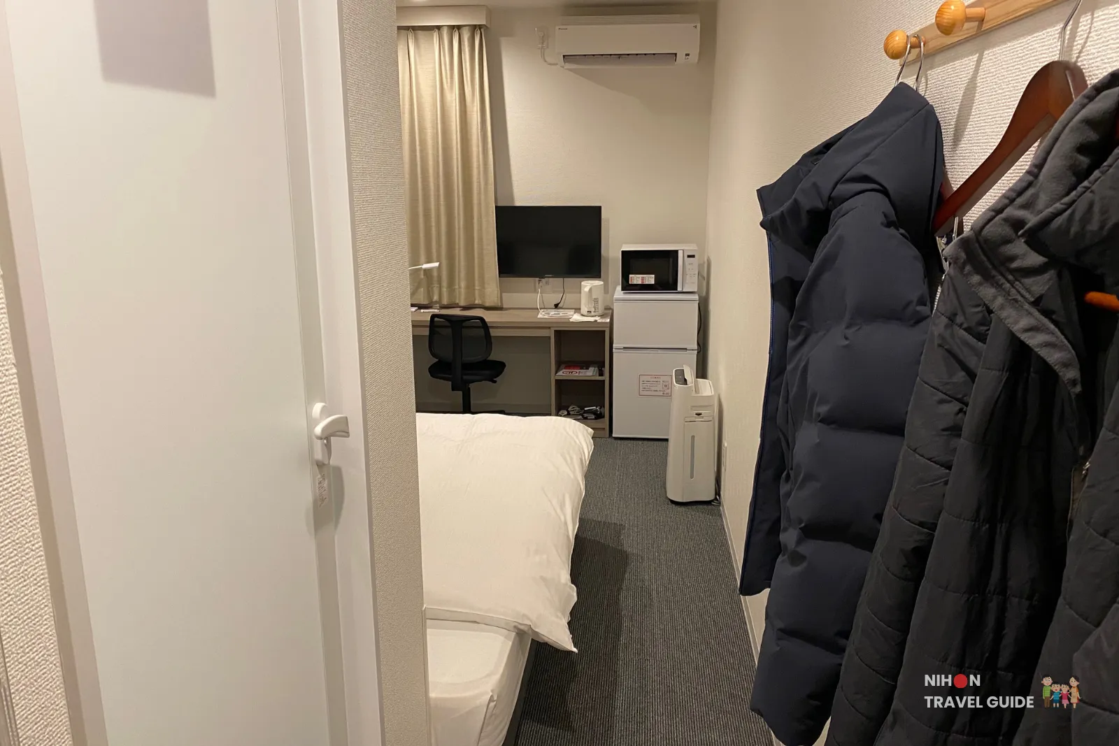 View from entrance corridor into Hotel R9 The Yard Tsukuba room showing bed, desk, TV, and outerwear hanging on hooks.