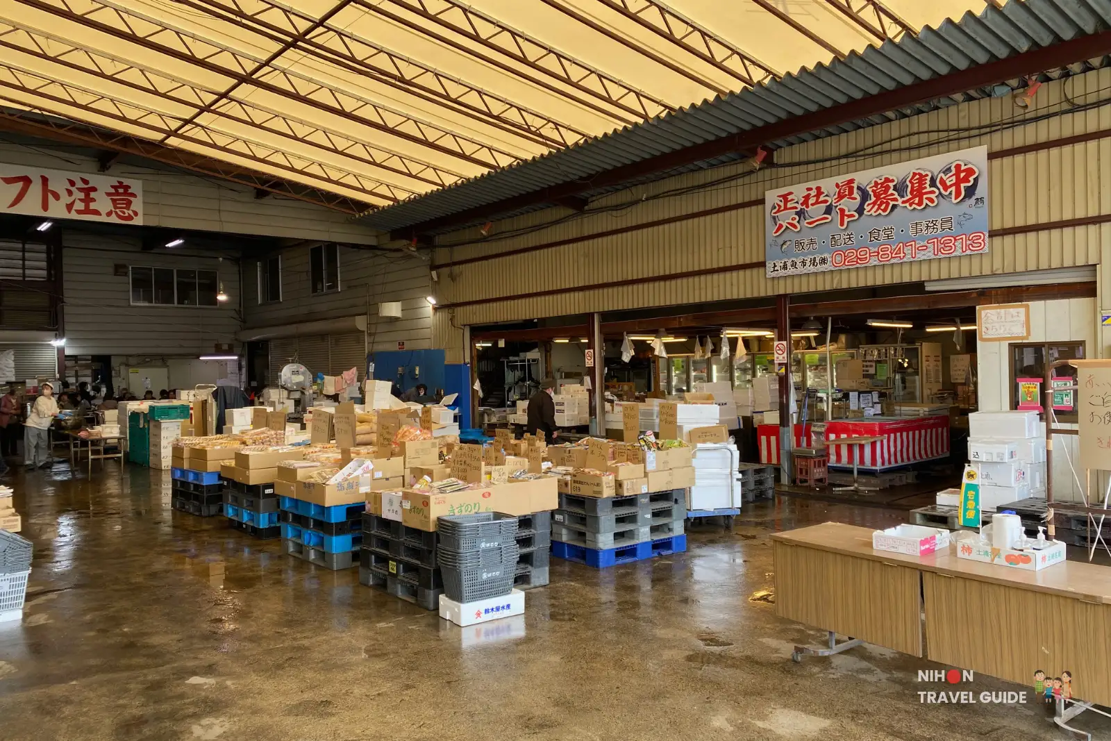 Busy interior of Tsuchiura Fish Market main wholesale hall with stacked produce crates, pallets of goods, forklift warning sign, shoppers and view of the fish stalls.
