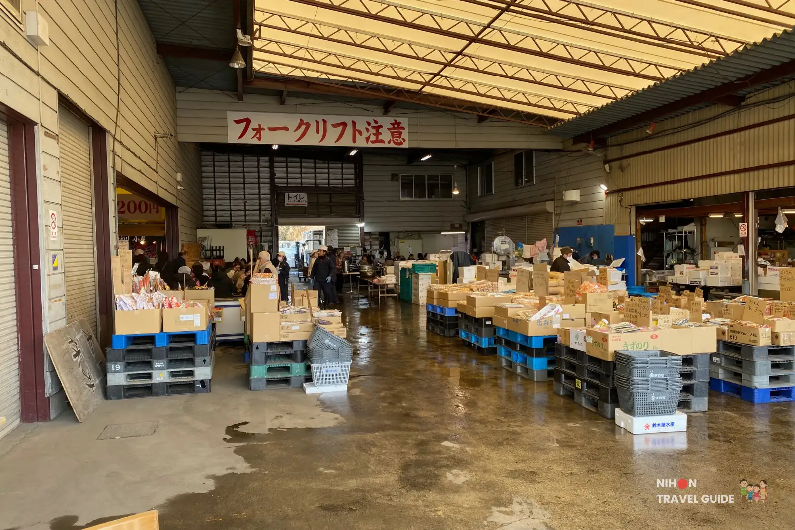 Busy interior of Tsuchiura Fish Market main wholesale hall with stacked produce crates, pallets of goods, forklift warning sign, and shoppers, authentic rural atmosphere.