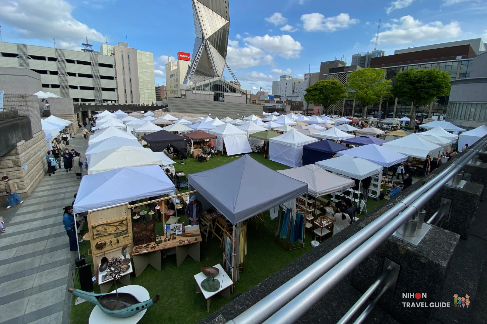 High-angle view of the expansive Aozora Arts & Craft Stall Market with dozens of white tents and shoppers on the lawn, Art Tower Mito towering in the background under a blue sky