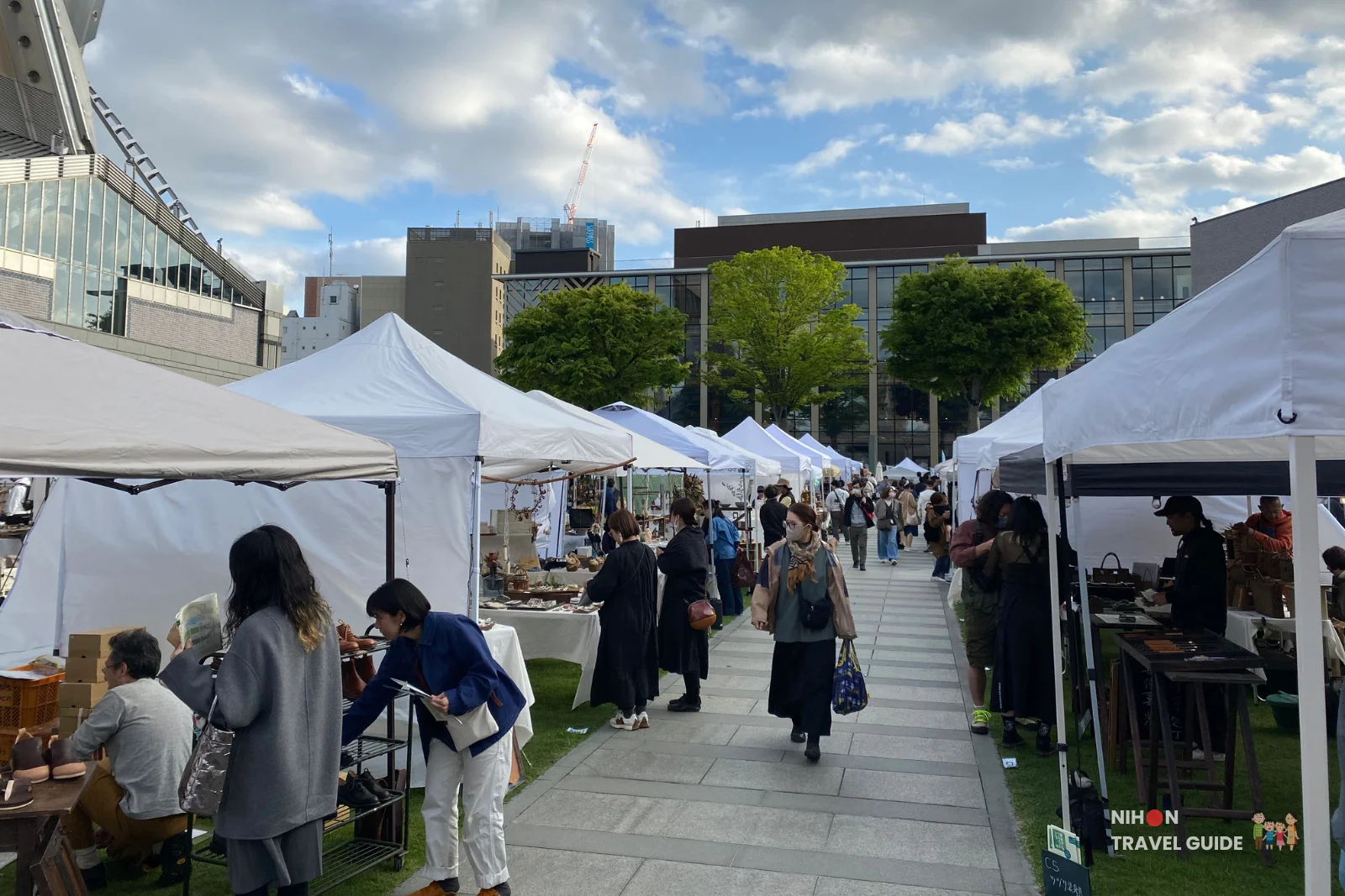 Crowded pathway lined with white vendor tents at the Aozora Arts & Craft Stall Market beside Art Tower Mito on a sunny day.