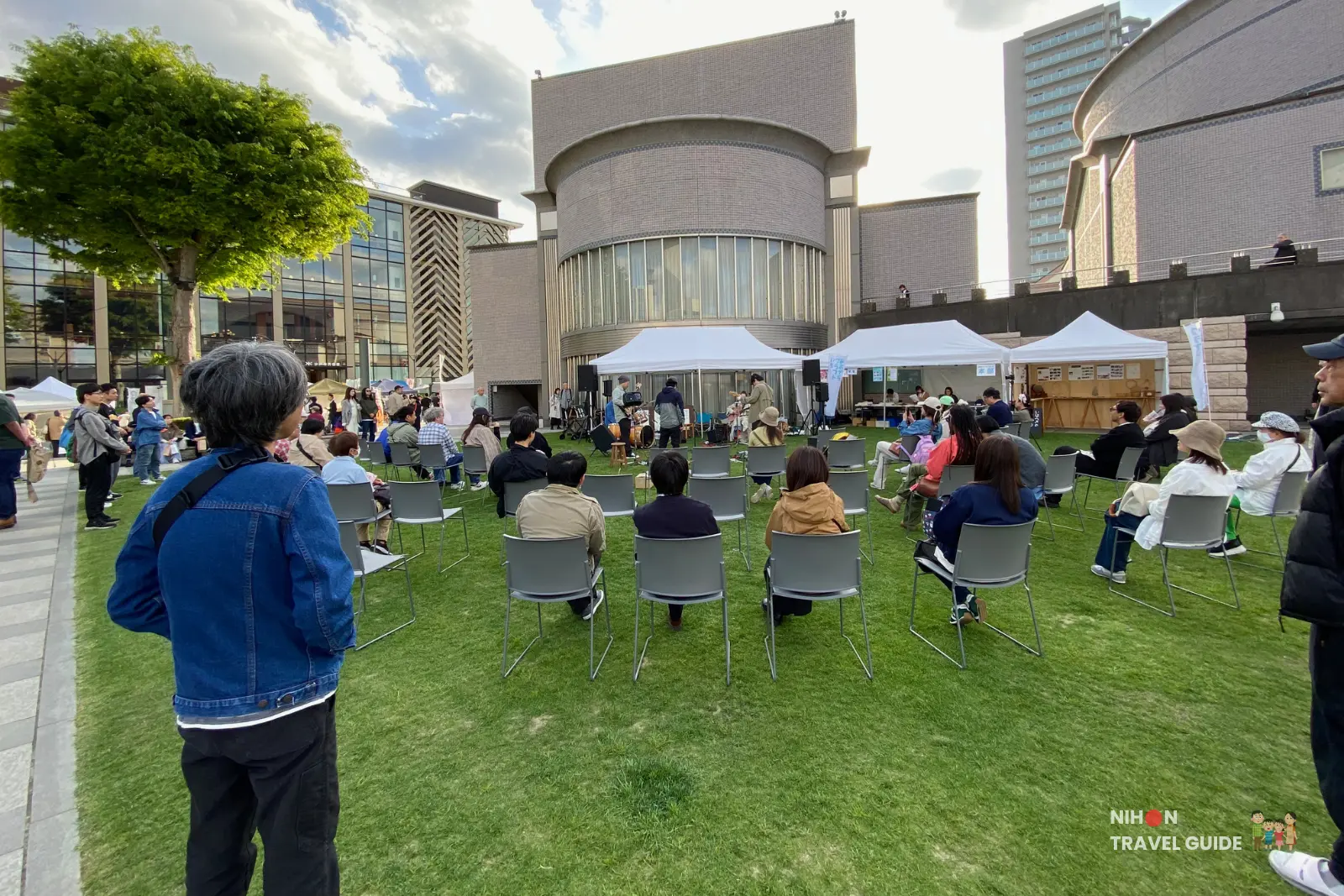 Audience seated on grey chairs on the lawn at Aozora Arts & Craft Market in front of Art Tower Mito, with event tents and performers in the background