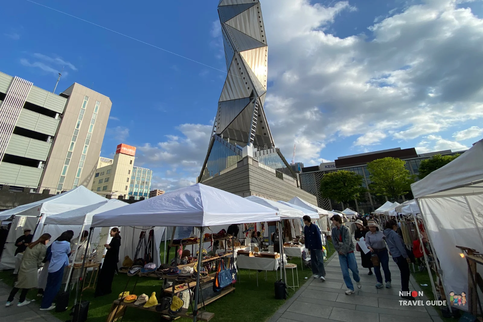 Visitors browsing handmade goods at outdoor stalls of the Aozora Arts & Craft Market with the striking Art Tower Mito visible in the background.