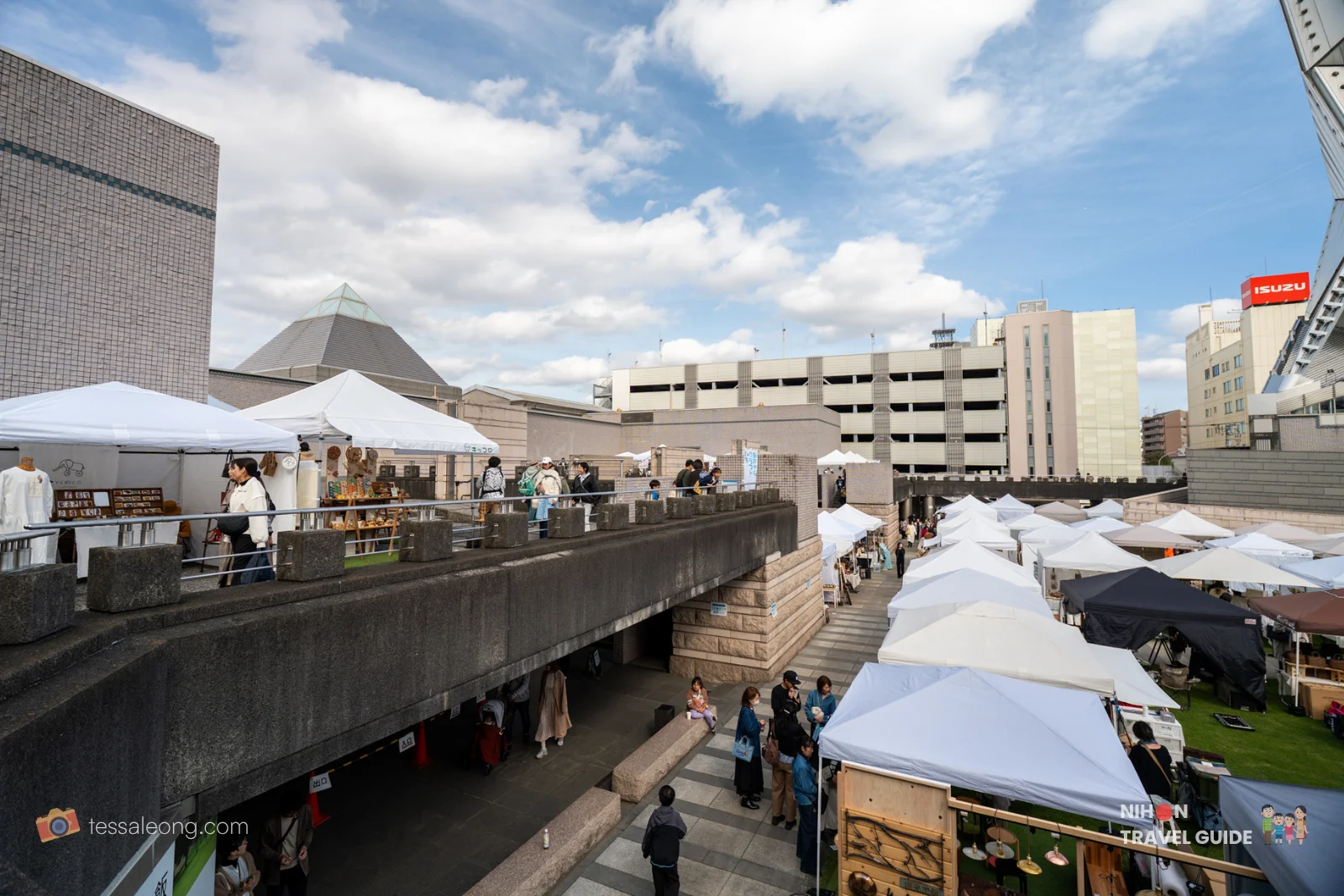 Wide view of the lively Aozora Craft Market with white tents, shoppers, and the cultural complex including the pyramid-shaped building at Art Tower Mito on a sunny day