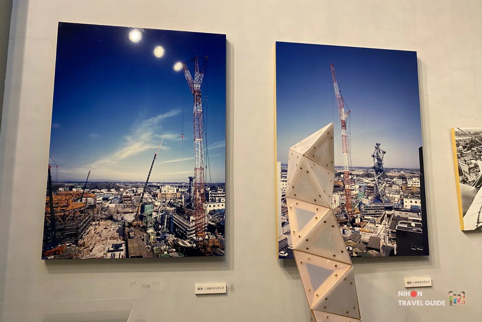 Framed exhibition photographs showing the construction of Art Tower Mito in March and April 1989, featuring large cranes against a blue sky and the early steel framework, displayed alongside a wooden tetrahedral tower model.