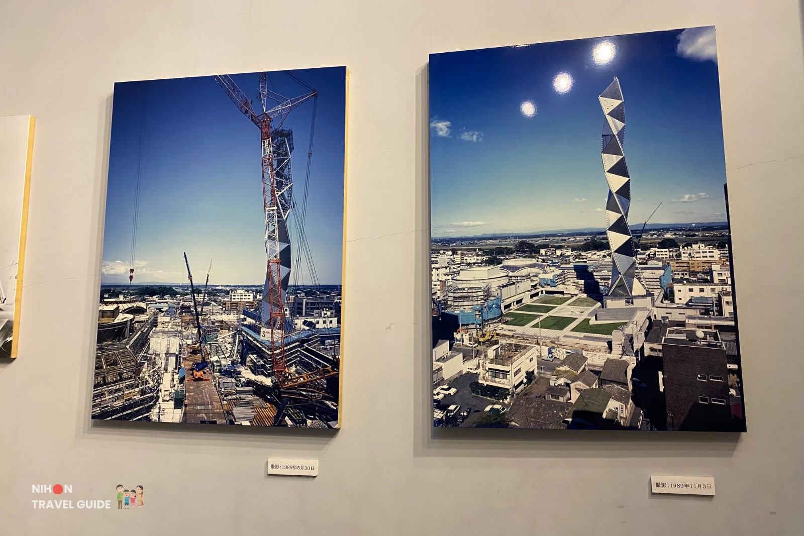 Two framed exhibition photographs showing the near-complete Art Tower Mito in May and November 1989, with the left image showing the titanium cladding being fitted and the right showing the finished spiral tower rising above Mito City