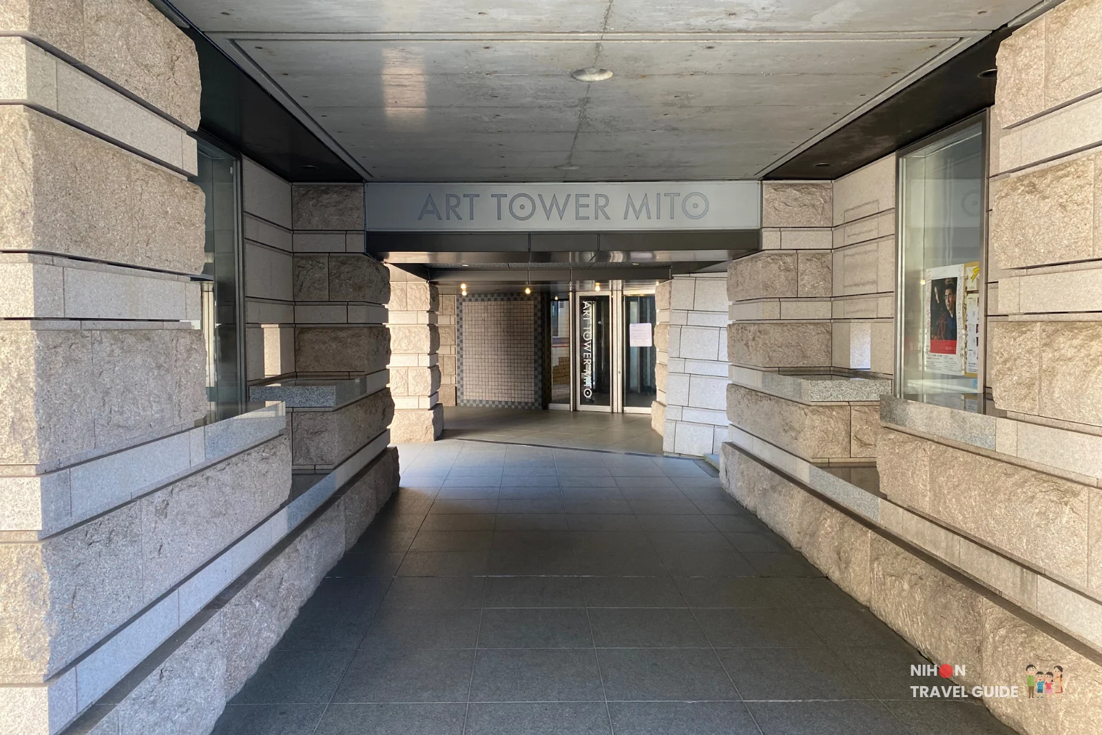 Covered stone-clad entrance walkway of Art Tower Mito with the building name displayed on a metal sign above the glass doors, flanked by granite walls and leading into the lobby, Mito City, Ibaraki, Japan