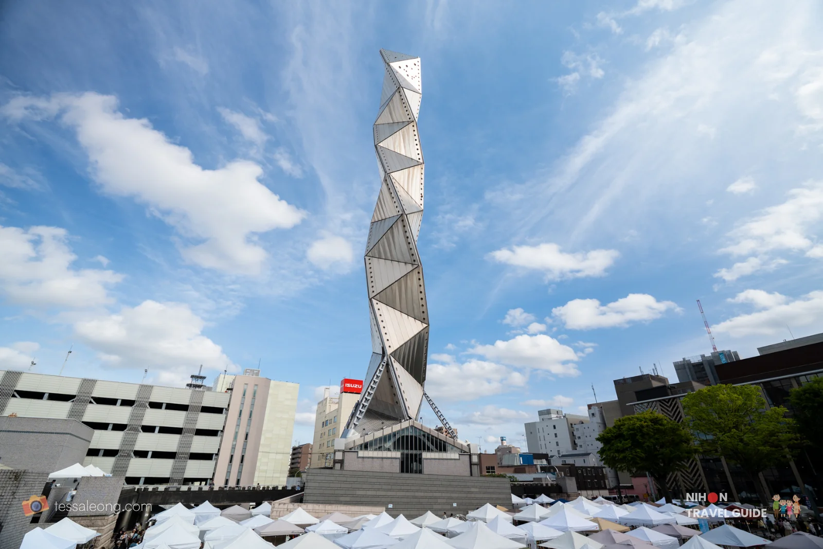 Low-angle view of Art Tower Mito's iconic 100m titanium spiral tower against a clear blue sky, with the Aozora Craft Market tents in the foreground in Mito, Ibaraki
