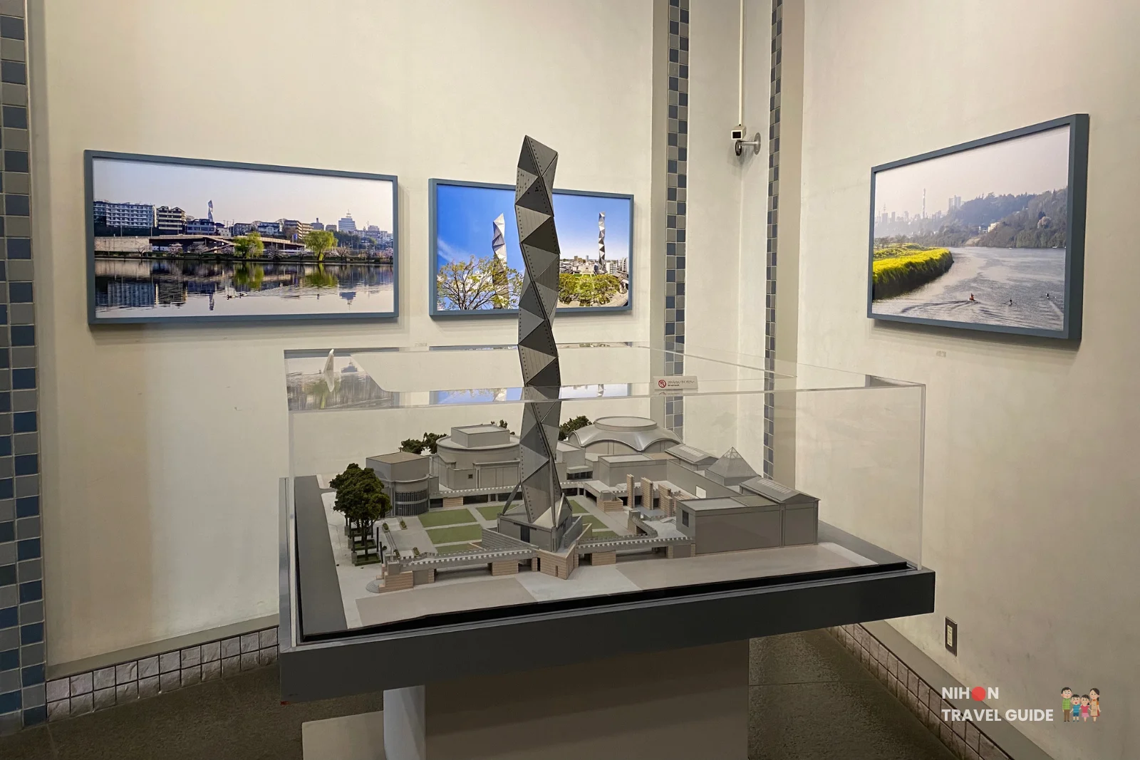 Detailed architectural scale model of the entire Art Tower Mito cultural complex displayed under a glass case, showing the spiral tower, concert hall, contemporary art museum, and surrounding plaza, with framed photographs of Mito City on the wall behind