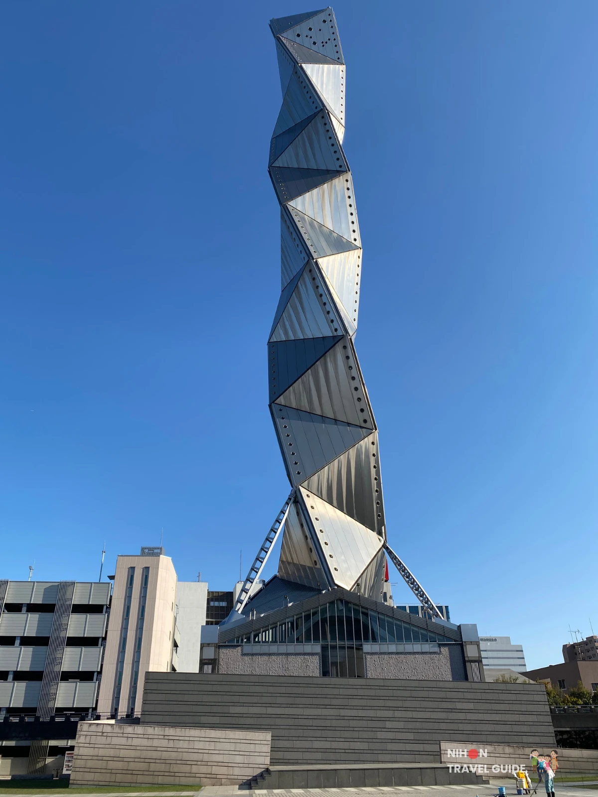 Full view of Art Tower Mito's 100-metre titanium-clad spiral tower photographed from the ground plaza, showing the complete triple-helix form made of 28 stacked tetrahedra with 57 triangular panels catching the sunlight against a clear blue sky, with visitors and surrounding buildings visible at the base, Mito City, Ibaraki, Japan