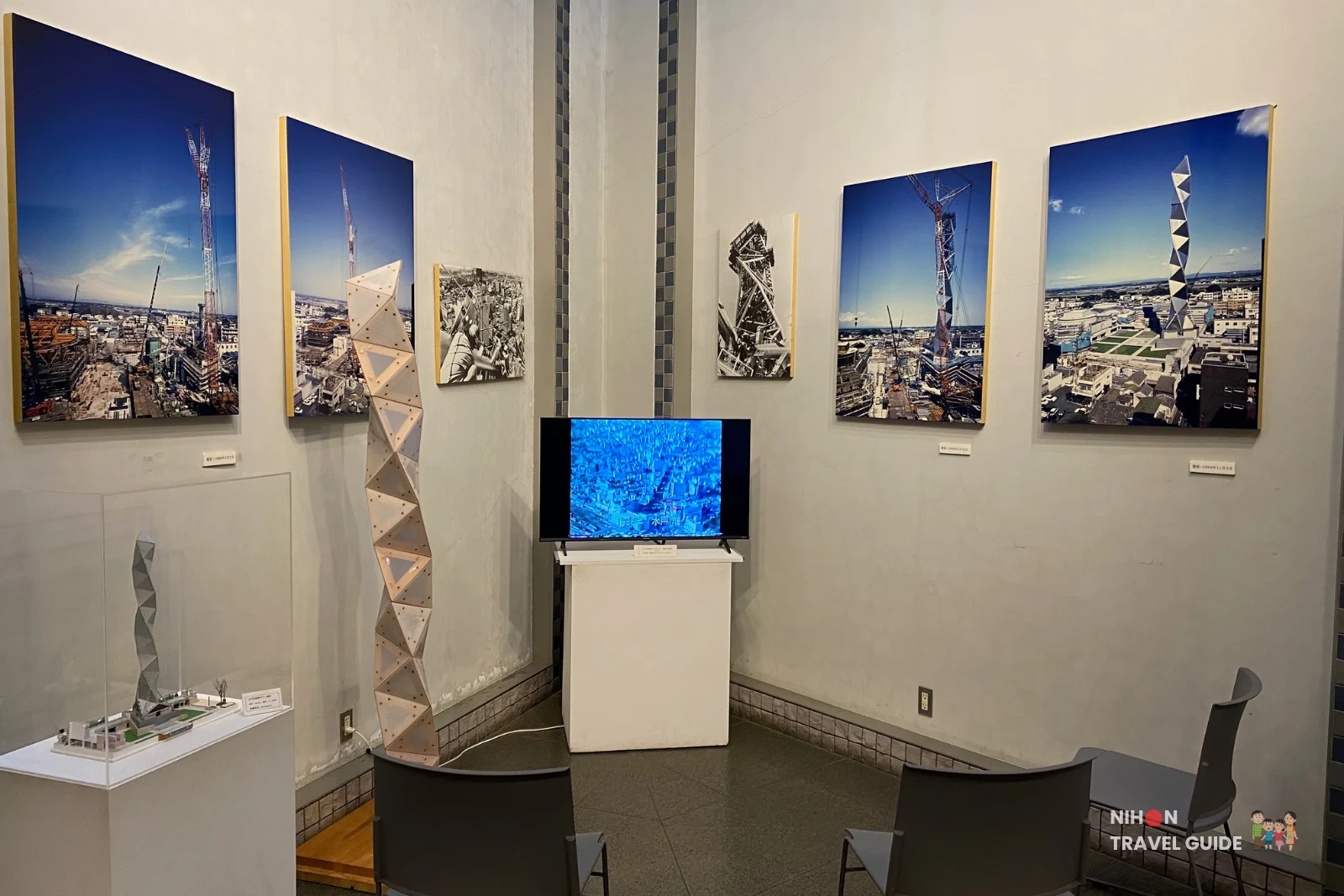 Ground floor exhibition room at Art Tower Mito displaying historical construction photographs, a wooden architectural model of the tower's tetrahedral structure, and a video screen showing an aerial view of Mito City, Ibaraki, Japan