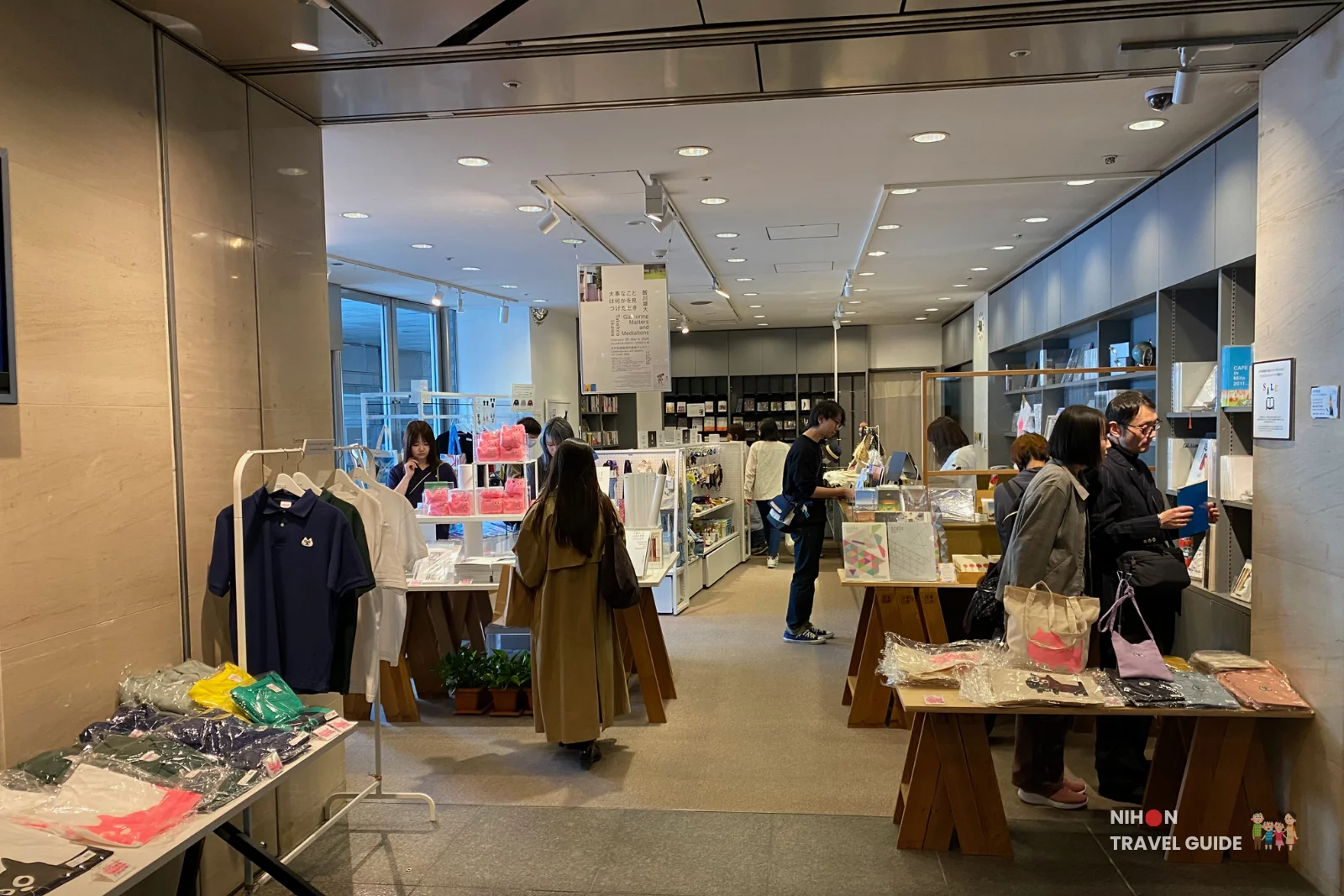 Busy museum gift shop inside Art Tower Mito with visitors browsing clothing, souvenirs, books, and handmade items on display tables and shelves.