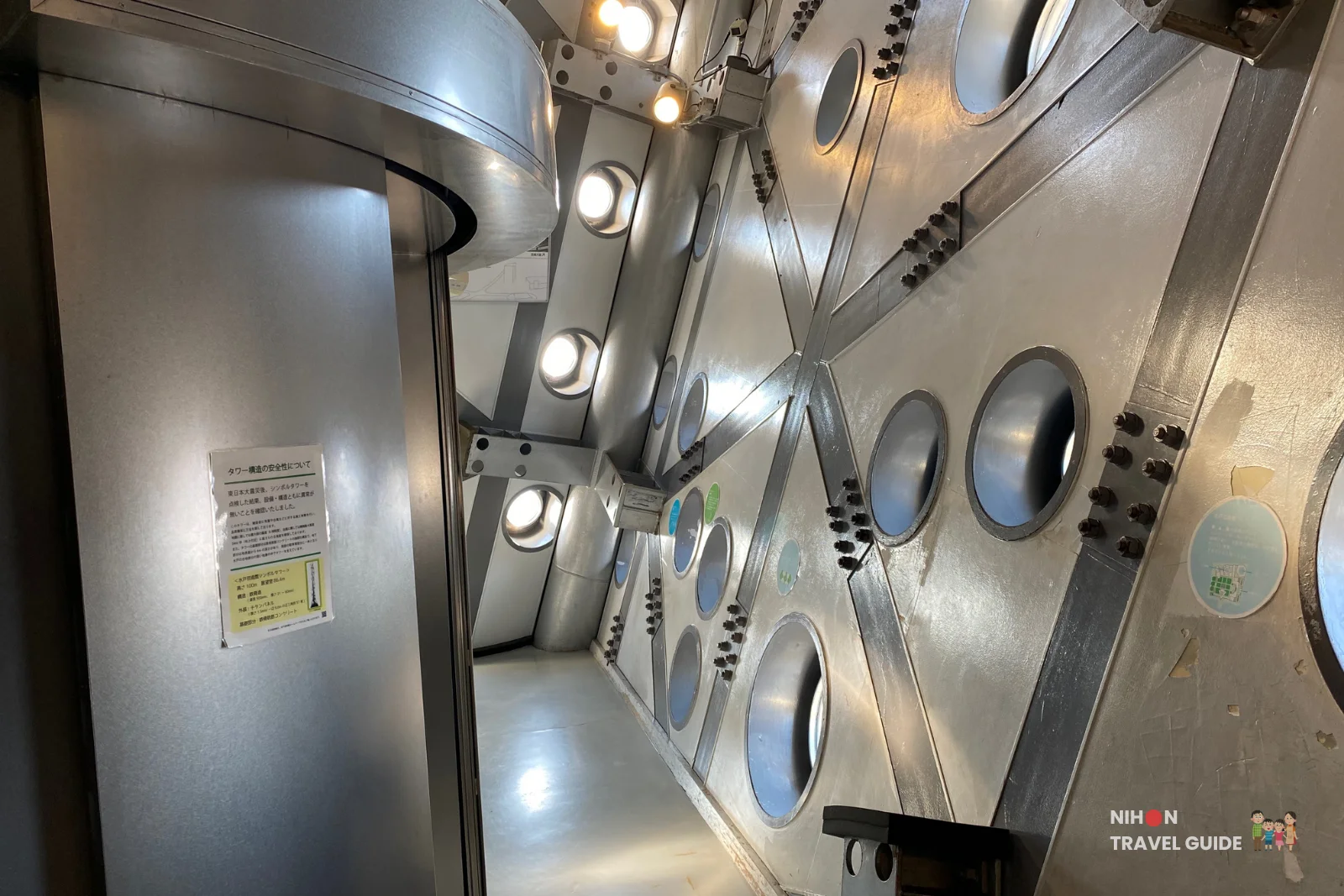 First view stepping out of the elevator onto the Art Tower Mito observation deck at 86.4 metres, showing the raw steel interior with angled cross-bracing, circular porthole windows of varying sizes set at different angles, and overhead spotlights, Mito City, Ibaraki, Japan