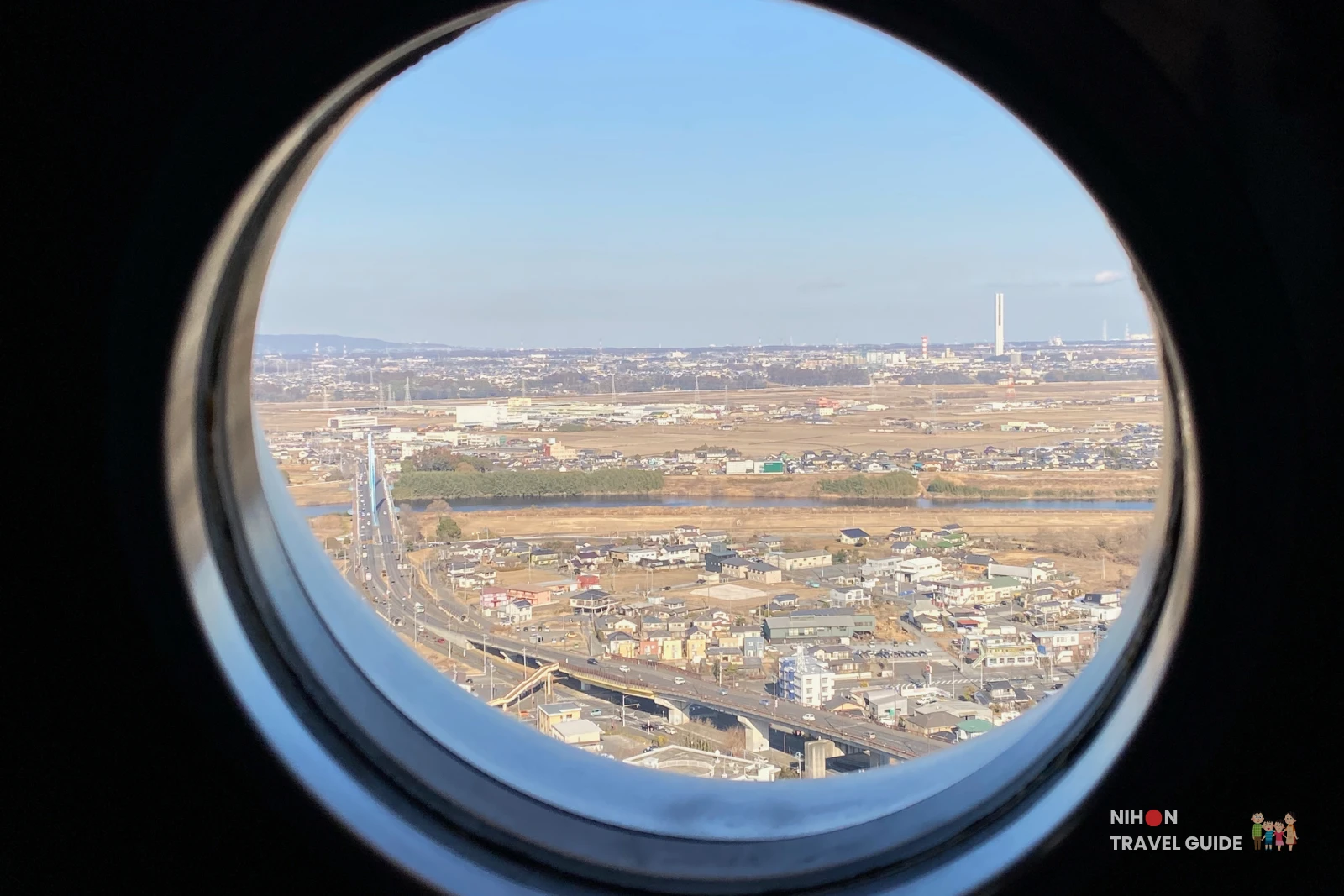 View through a circular porthole window on the Art Tower Mito observation deck at 86.4 metres, looking north-east toward the Bandai Bridge (cable-stayed bridge with blue pylon) spanning the Naka River, with the flat Ibaraki plains stretching to the horizon and the distant chimneys of the G1 Tower area (Hitachi elevator research tower) faintly visible on the right, Mito City, Ibaraki, Japan