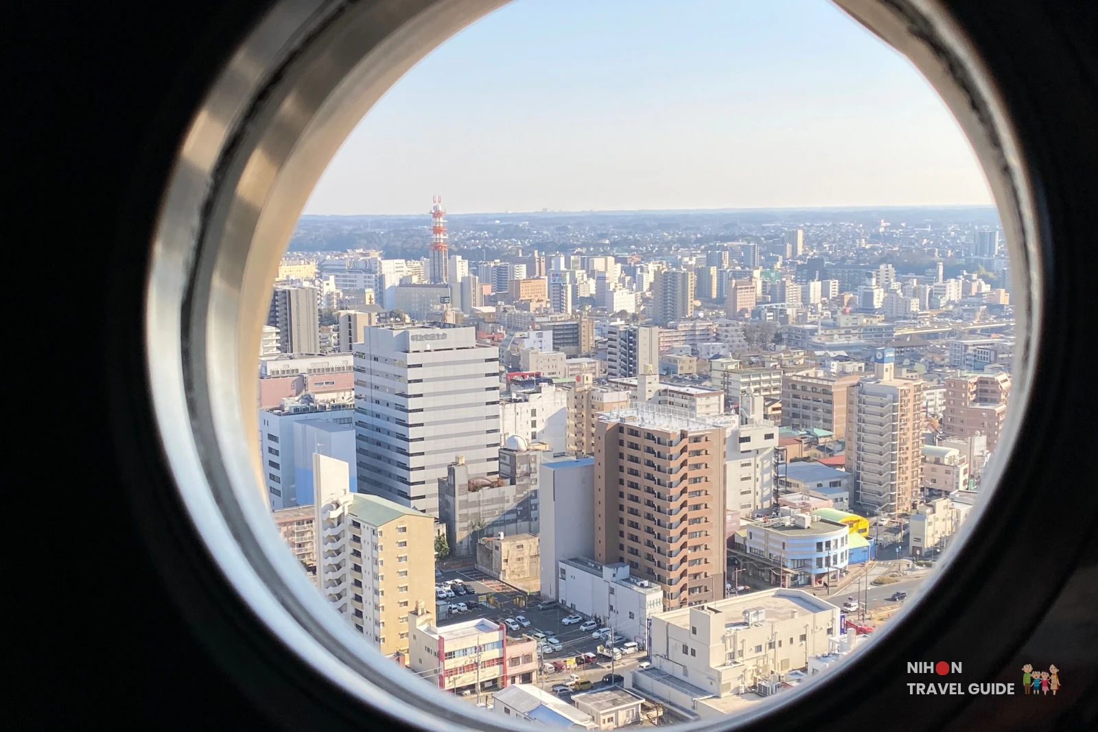 View through a circular porthole window on the Art Tower Mito observation deck looking east over Mito City's dense urban landscape toward the Pacific Ocean coast at Oarai approximately 12km away, with a red-and-white broadcast communications tower visible on the horizon, Ibaraki, Japan