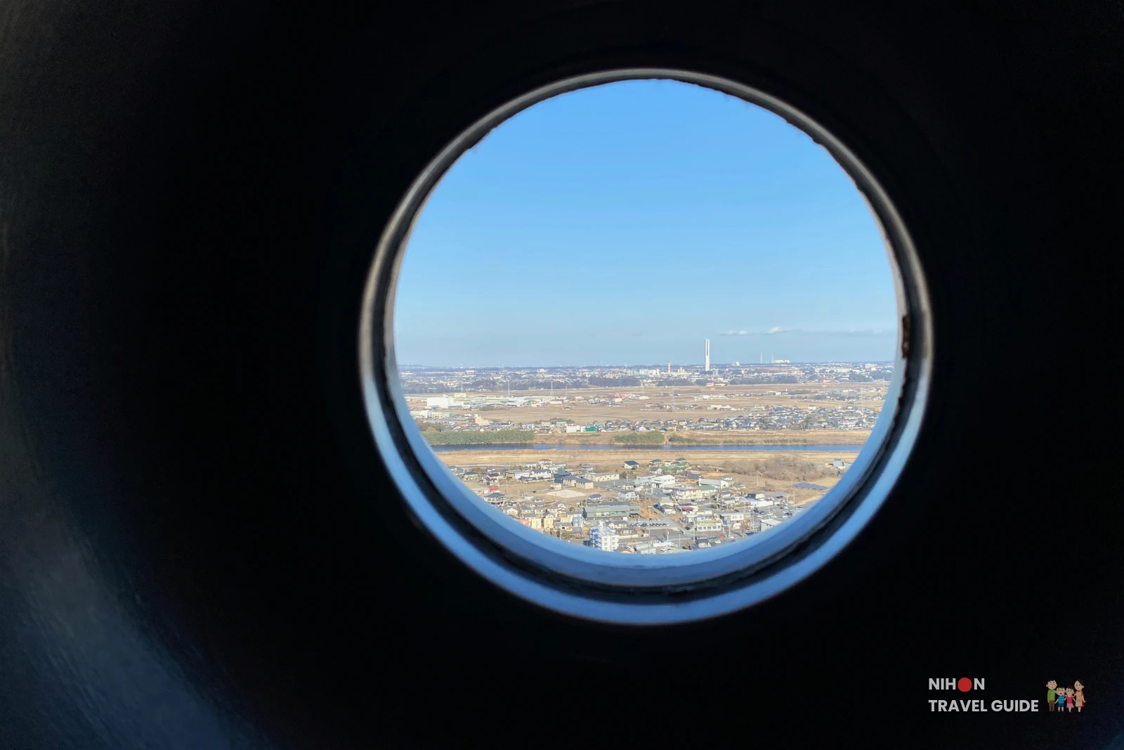 View through a circular porthole window on the Art Tower Mito observation deck at 86.4 metres, showing the Hitachi G1 Tower — Japan's tallest elevator research tower at 213.5 metres — visible as a slim white needle rising above the flat Ibaraki plains on the horizon, with the Naka River and residential suburbs of Mito City in the foreground, Ibaraki, Japan