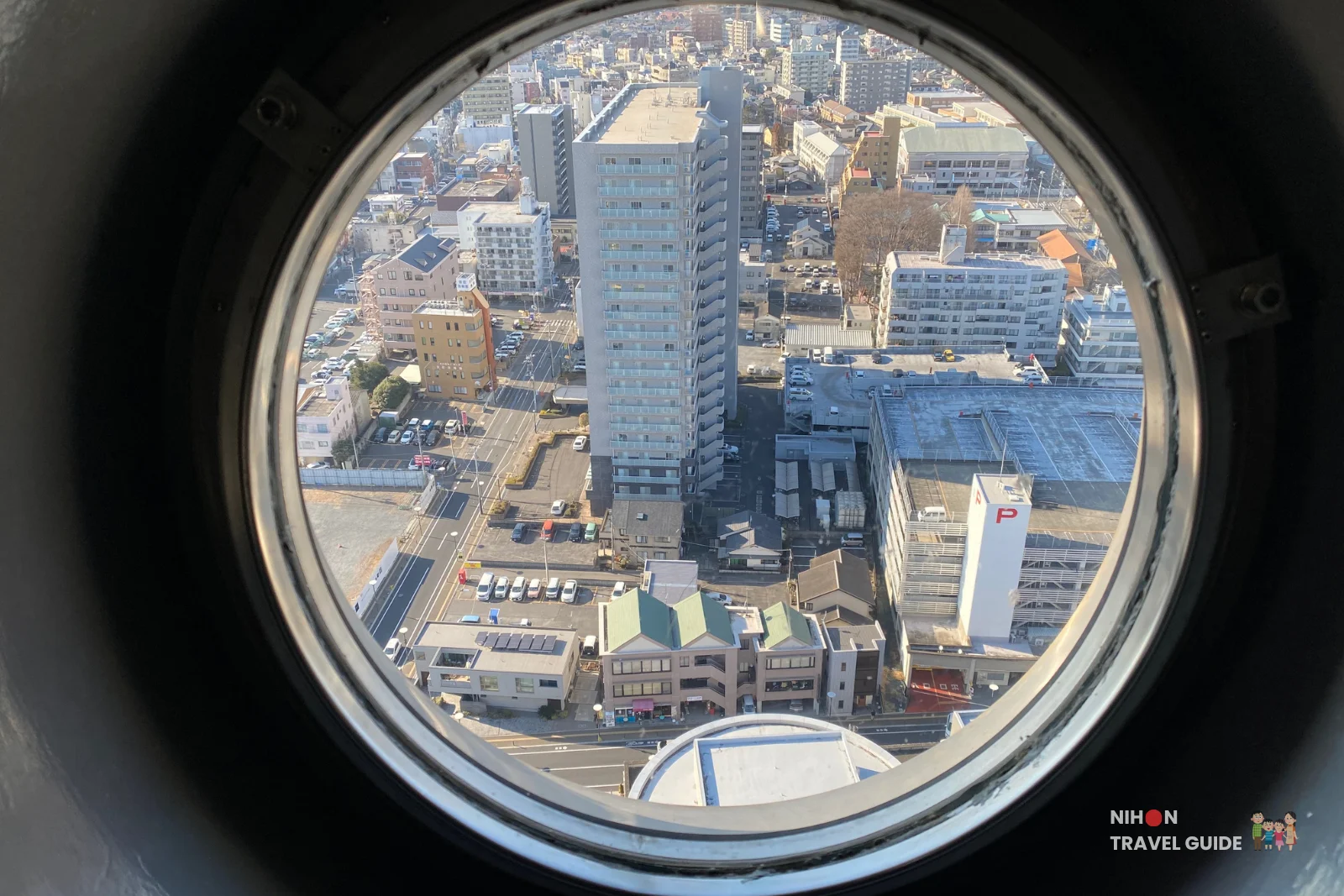 Bird's-eye view of Mito City centre through a circular porthole window on the Art Tower Mito observation deck at 86.4 metres, looking directly down onto the surrounding urban streets, parking lots, and buildings including a tall glass residential or hotel tower, with the dome roof of the Art Tower Mito concert hall visible at the bottom of the frame, Mito City, Ibaraki, Japan